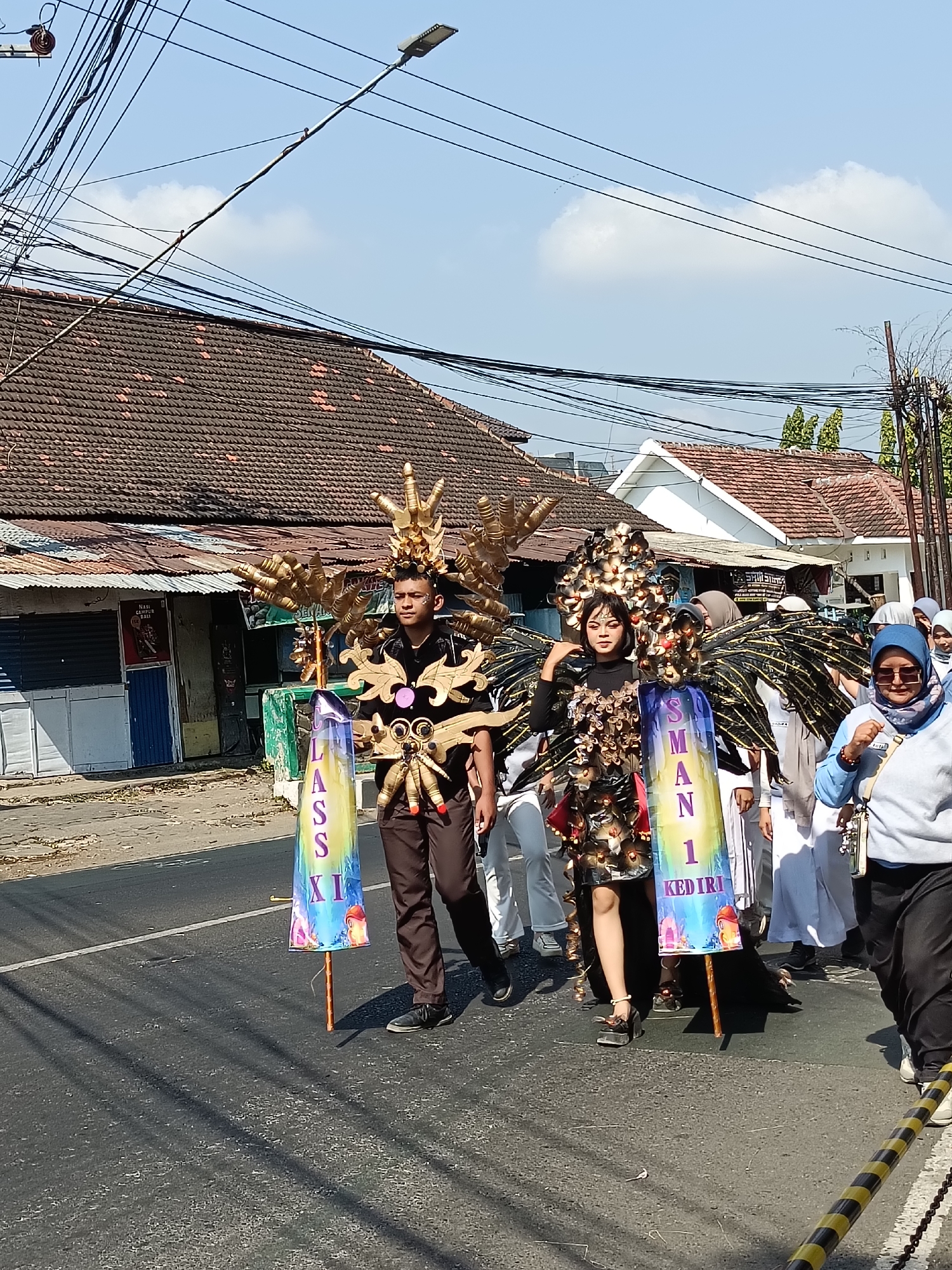 In Kediri City, Indonesia, under a clear blue sky with scattered clouds, a parade is underway on a paved road lined with buildings. Two individuals, a male and a female, are at the forefront, dressed in elaborate, golden and black winged costumes with ornate headpieces. The male is wearing brown pants and a black shirt, while the female is in a black dress and high heels. They are holding tall, cylindrical banners. The male's banner is multicolored with text that appears to read "LASSI XI". The female's banner is also multicolored with text that appears to read "SMAN 1 KEDIRI". Behind them, a group of people in white attire, some wearing headscarves, are walking in the parade. The buildings on the left of the street have tiled roofs and some appear to be shops. Power lines crisscross the sky, and a street lamp is visible. The road surface shows shadows and some debris.