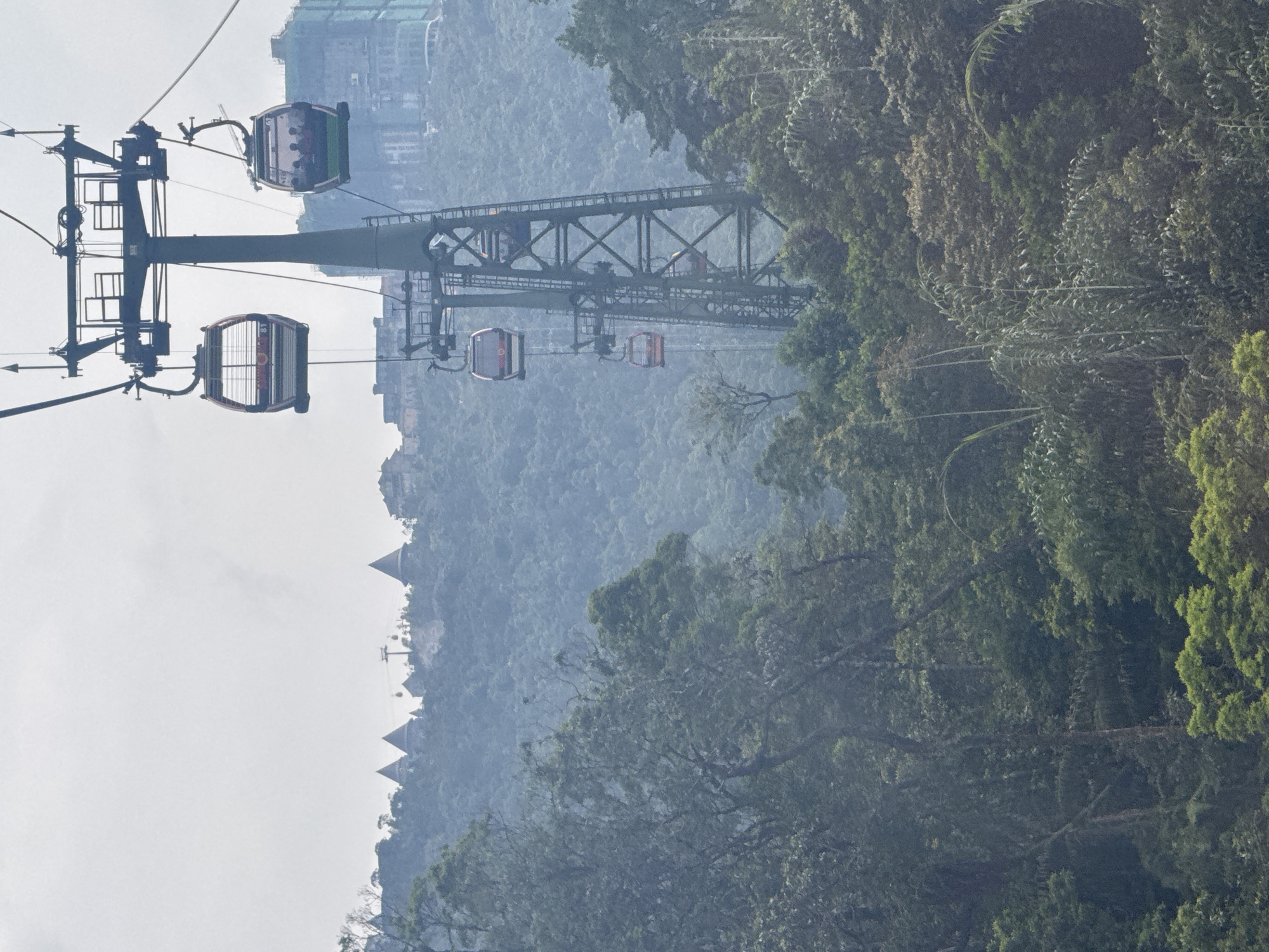 A cable car system traverses a verdant, mountainous landscape in Đà Nẵng, Vietnam. The main subject is a prominent dark green support tower, from which multiple cable car gondolas are suspended. Two gondolas are clearly visible in the foreground, with several others receding into the distance along the cable lines. The gondolas are modern, enclosed cabins designed for passenger transport.
The scene is set against a backdrop of dense, forested mountains. Lush green trees, varying in shade and texture, cover the slopes, from the immediate foreground to the hazy distant peaks. On a distant mountain ridge, a series of structures with distinctive conical or pointed roofs can be discerned, suggesting an architectural style common to resort areas or attractions in the region, such as Bà Nà Hills.
The weather appears hazy or overcast, diffusing the daylight and creating a soft, somewhat muted palette, especially in the background. The cable cars are either in motion or poised for operation, indicating an active transportation system connecting different parts of the mountainous terrain. No identifiable human figures are clearly visible within the gondolas due to the distance and resolution. No legible text is visible on any of the objects. The image conveys a sense of expansive natural scenery enhanced by modern infrastructure.