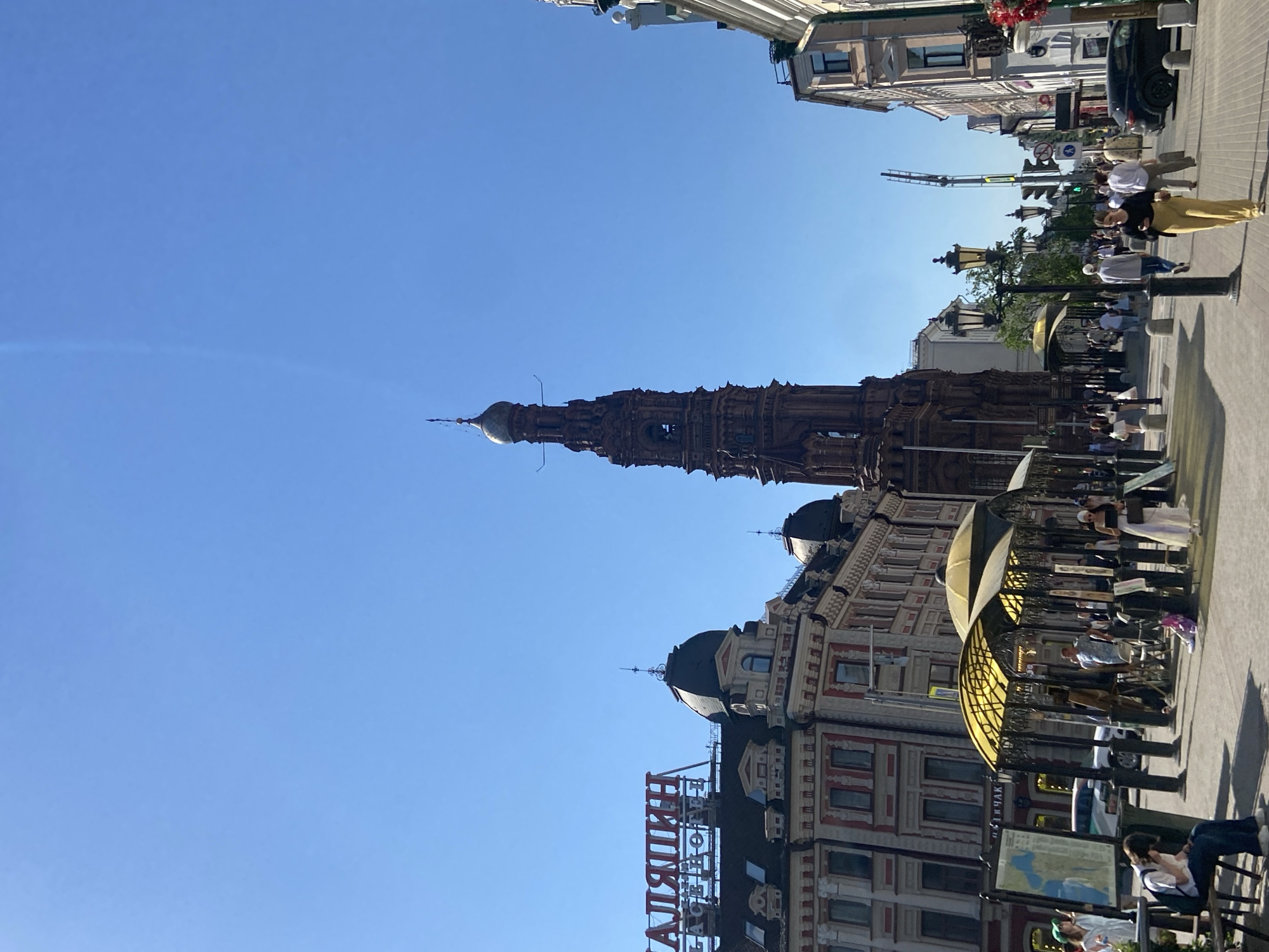 The image depicts a street scene in Kazan, Russia, featuring the leaning Suumbike Tower of the Kazan Kremlin. The tower, of reddish-brown brick construction, dominates the center of the frame against a clear blue sky. To the right of the tower, pedestrians walk along a paved sidewalk. Further to the right, people are seated at outdoor tables under awnings. Architectural details of adjacent buildings are visible, including windows, facades, and decorative elements. A signage with Cyrillic letters is also visible, and a black car parked along the street. The orientation of the photo is rotated.