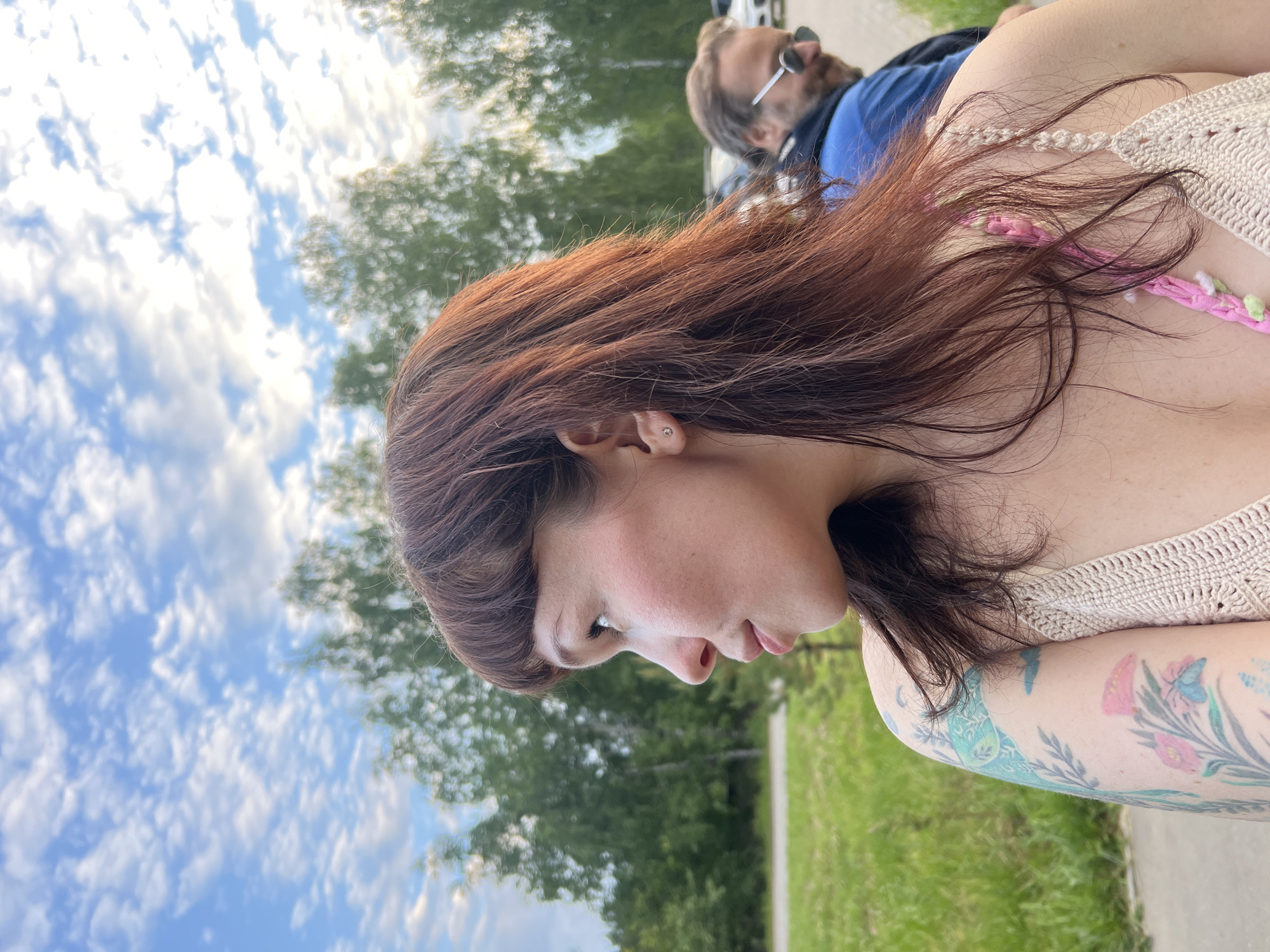 An outdoor, medium shot captures a woman with auburn hair and bangs, wearing a knitted top in shades of beige and pink. She is turned to her left, with her profile visible against a backdrop of trees and a cloudy sky. Her left arm, visible in the foreground, features a tattoo of flowers and a butterfly. Behind her, a man with sunglasses and a blue jacket is partially visible. The scene appears to be in a park or outdoor recreational area, possibly in the city of Chaikovsky, Russia, given the prevalence of birch trees and the cloudy sky.
