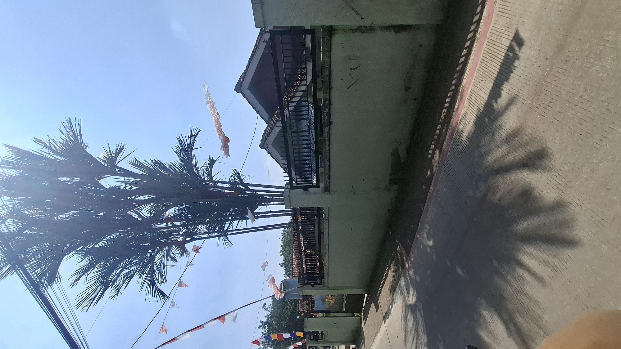 A sunlit outdoor scene depicts a low-rise building with a tiled roof, surrounded by a pale green painted wall and a black metal fence. Several palm trees are visible behind the fence, their fronds extending towards a bright blue sky. A string of red and white triangular flags, likely national flags, is strung across the scene, along with longer white and red striped banners. The ground in the foreground is paved and shows the distinct shadow patterns of palm fronds. The location is identified as Ciharashas, Indonesia.