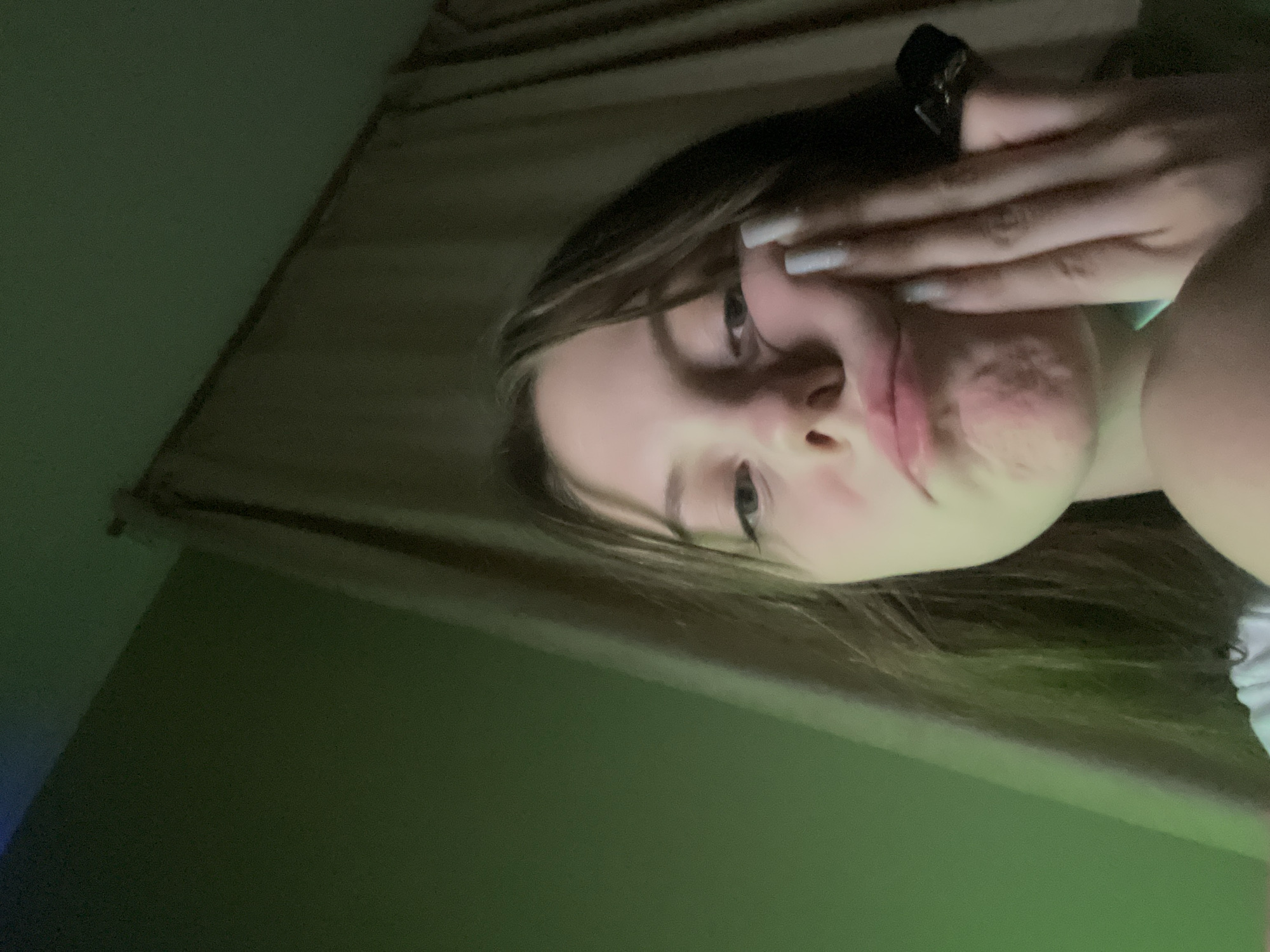 A close-up, indoor photograph shows a young woman holding her hand to her face. She has fair skin, light brown hair, and appears to be wearing light nail polish. A dark object, possibly a handheld device, is visible above her hand. The background includes a wall and a curtained window. The scene is illuminated by green light. There is no information present in this image about location.