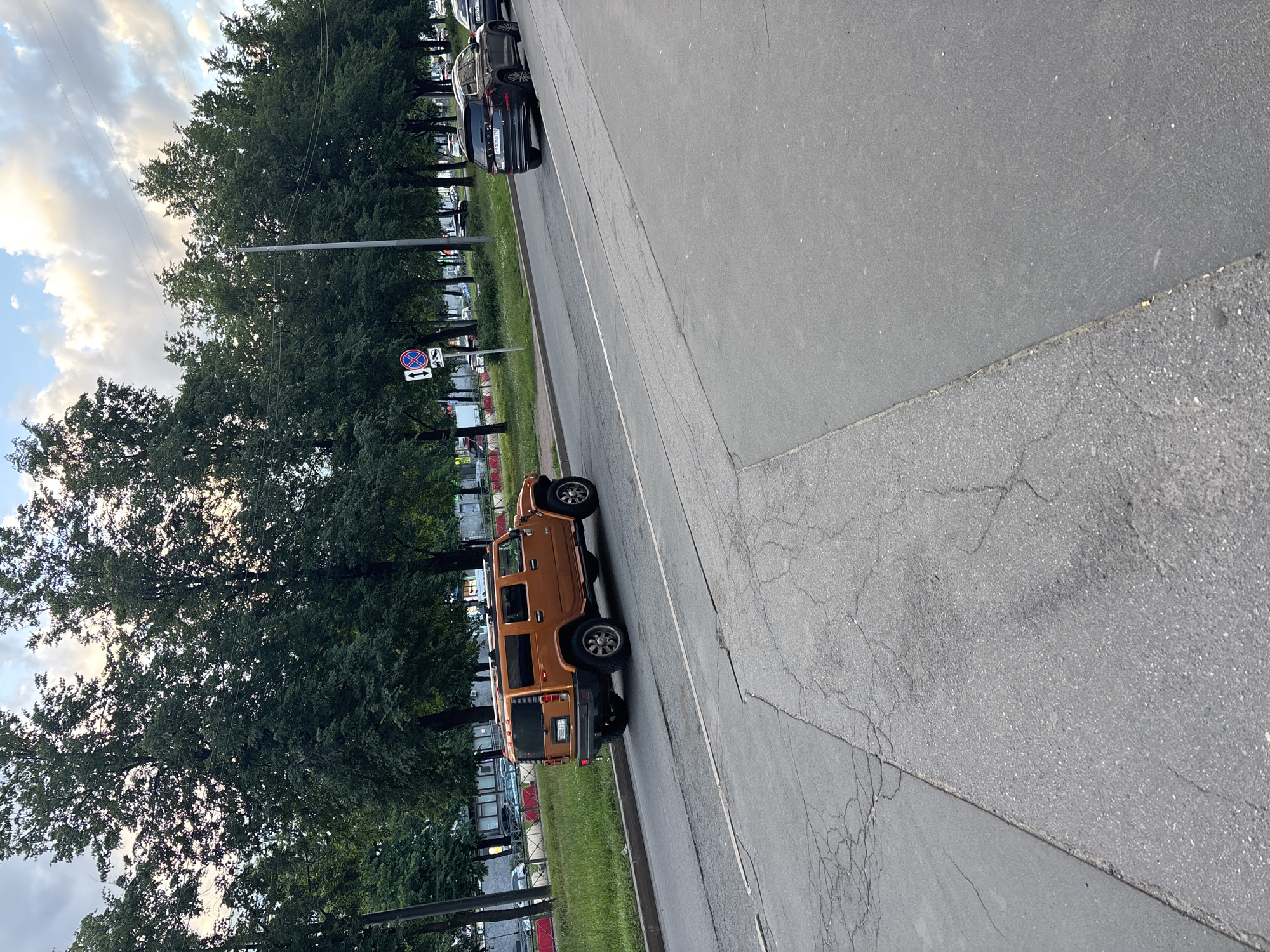 An orange Hummer H2 SUV is parked on the right side of a paved road. The SUV is facing left, with its rear to the viewer. The vehicle has a black license plate with white lettering that is not legible. The road is gray asphalt and appears cracked in places. To the left of the road is a grassy area with several tall trees. In the background, there are more trees and some buildings. The sky is partly cloudy with a mix of blue and white. Another black SUV is parked on the right side of the road, further in the distance.