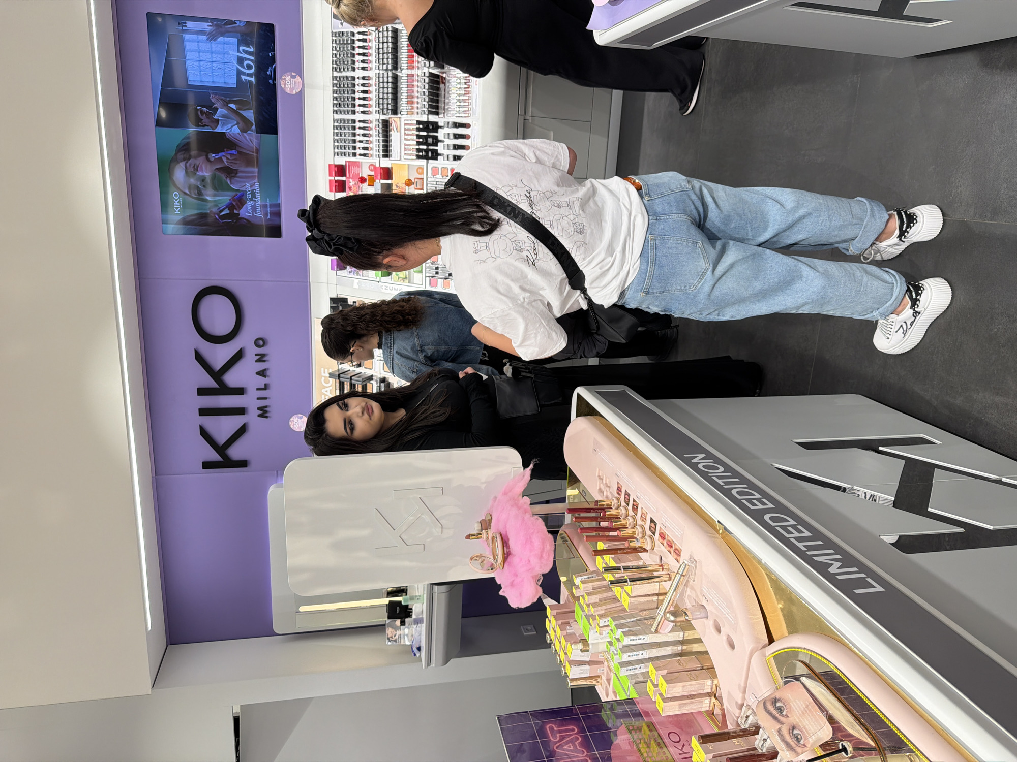 The image shows the interior of a Kiko Milano store, possibly in Burgas, Bulgaria. A purple wall with the white "KIKO MILANO" logo is visible. A video screen is affixed above the logo. Three people are observed shopping: one is wearing a white t-shirt, light blue jeans, and white platform sneakers; another is wearing a black jacket, and the third is wearing a denim jacket. Makeup products are displayed on shelves labeled "LIMITED EDITION".