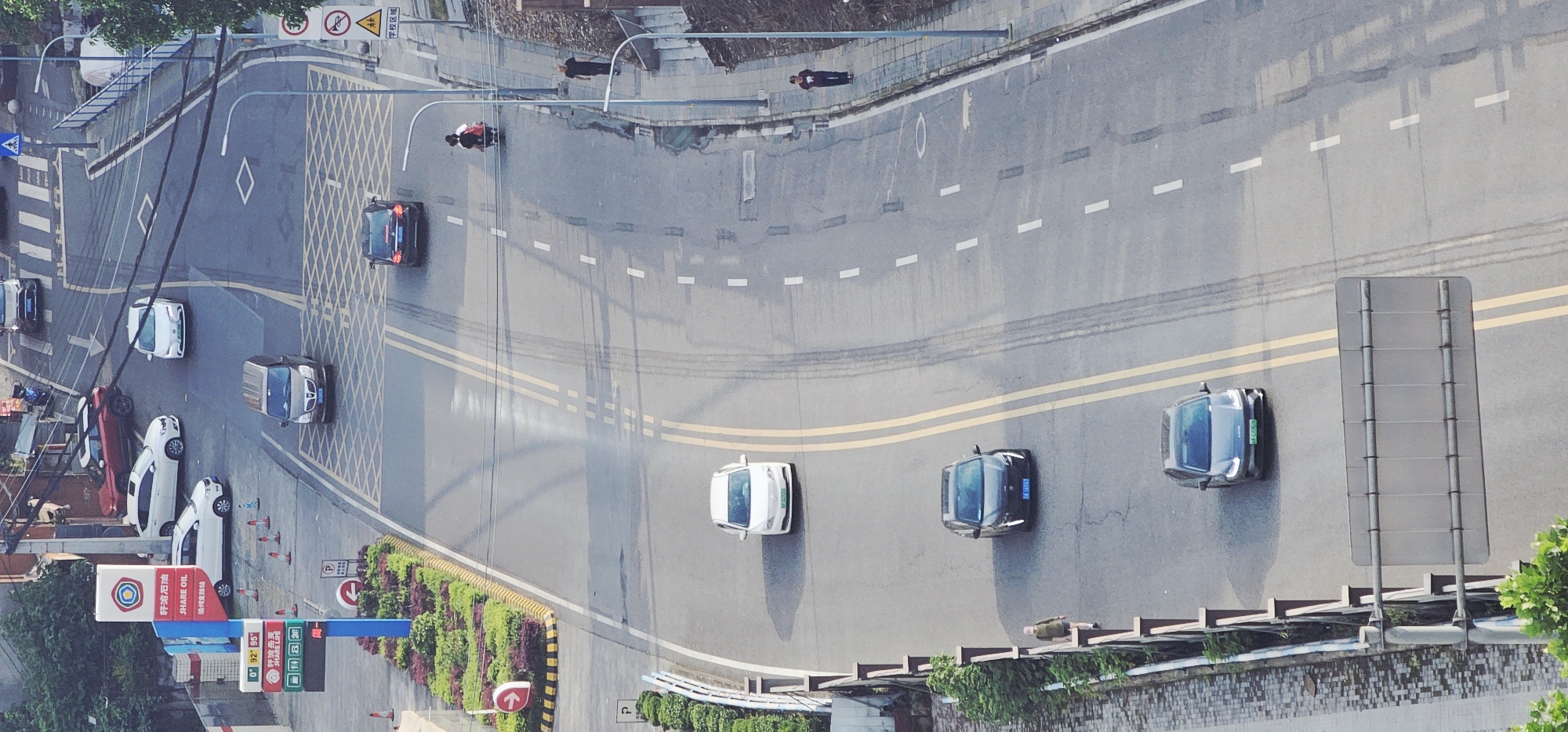 The image is an aerial shot of a street in Jiulongpo District, China. The street is paved and has multiple lanes with dashed white lines separating them, as well as solid double yellow lines in the center. There are several cars on the street, some parked and some driving. A person on a motorbike is visible at a road junction. There is a gas station located to the left with associated signage. There is greenery in the form of bushes and trees along the sides of the road. There is a pedestrian walkway on the right-hand side. Streetlights are visible. Road markings and street signs are also visible. The scene appears to be a typical urban setting with vehicular and pedestrian traffic.