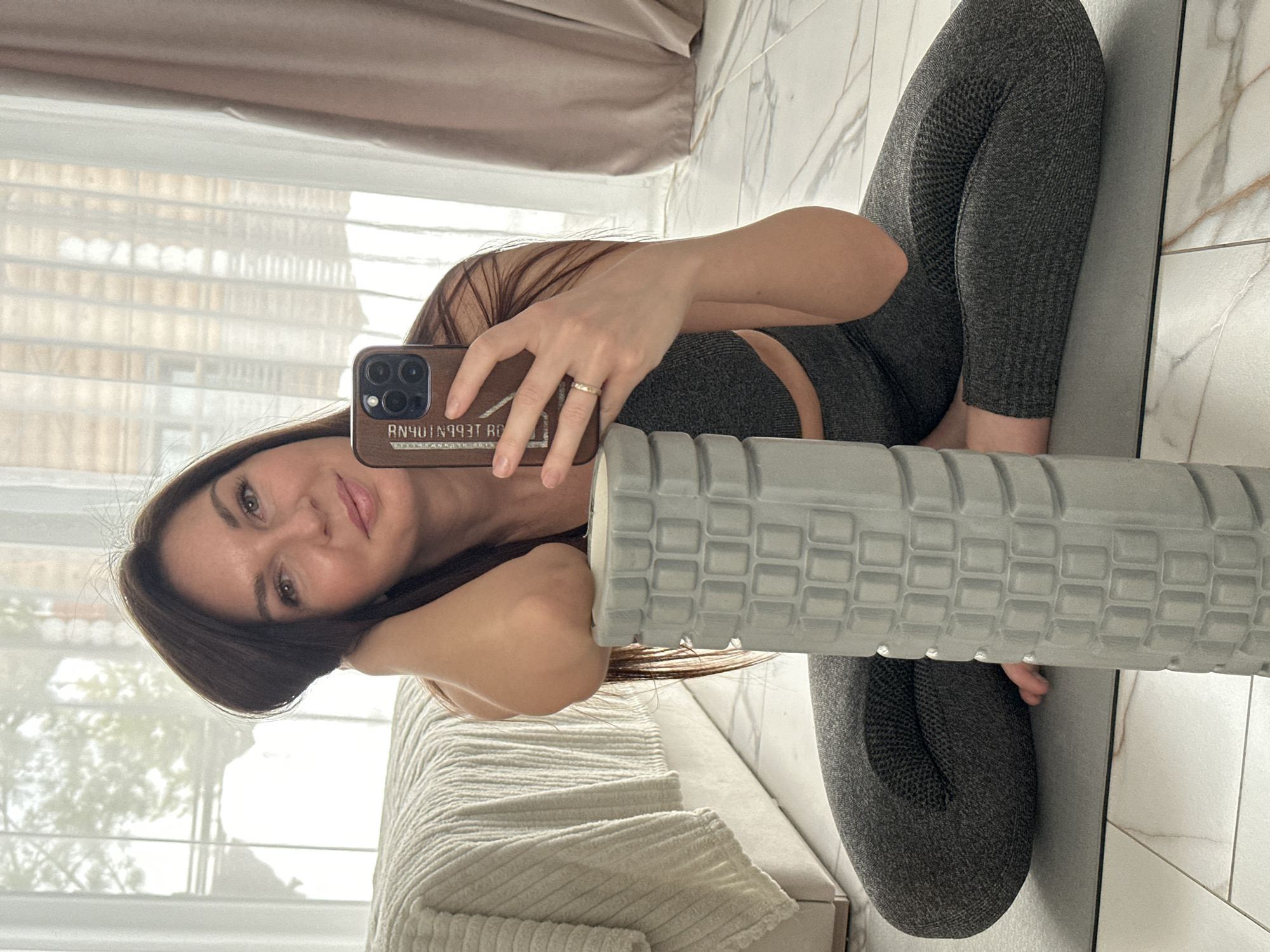 An adult female is depicted taking a selfie indoors, presumed to be in Chaikovsky, Russia. She is seated on a grey yoga mat on a light-colored marble or tiled floor, dressed in a dark grey textured two-piece athletic set consisting of a sports bra and leggings. Her dark brown hair is long. She holds a smartphone with a dark case, displaying three rear camera lenses, in her right hand. The phone case shows reversed text, "Я ВЫБИРАЮ ЖИЗНЬ" (I CHOOSE LIFE). Her left forearm rests on a grey, textured cylindrical foam roller positioned vertically in front of her. A light-colored ring is visible on her left ring finger. She is looking directly at the camera with a slight smile. In the background, a bed with a light-colored, textured duvet is visible to the left. Large windows covered by sheer white curtains allow diffused natural light into the room. Beyond the windows, a blurred exterior shows trees and potential snow cover, with what appears to be part of a wooden building structure. Light brown curtains are drawn to the right of the windows.