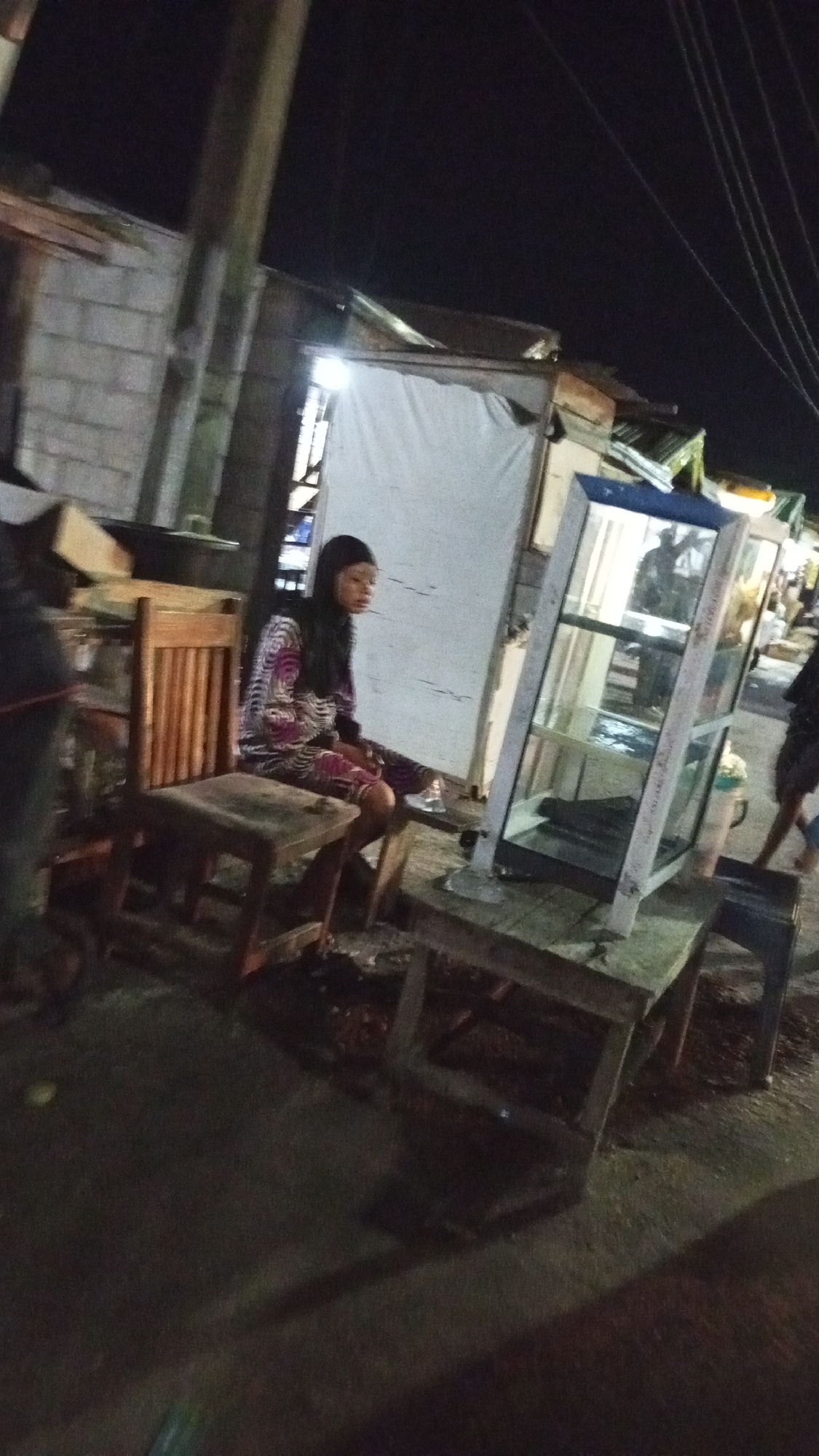The image, taken at night in what appears to be a low-lit area of Adankolo, Nigeria, shows a woman seated on a wooden stool at a makeshift outdoor setup. She is wearing a patterned long-sleeved shirt or dress and has dark hair. In front of her is a wooden table on which sits a glass display case. Another wooden chair is beside her. Behind her, a white sheet or fabric is hung up, possibly as a backdrop. To the left, there are stacked items and building materials, suggesting a somewhat cluttered environment. There is an unfinished concrete wall to the left. Overhead power lines are visible in the background. The ground appears to be paved but uneven. People are vaguely visible in the background. The scene suggests an informal commercial activity or a temporary setup in an outdoor space.