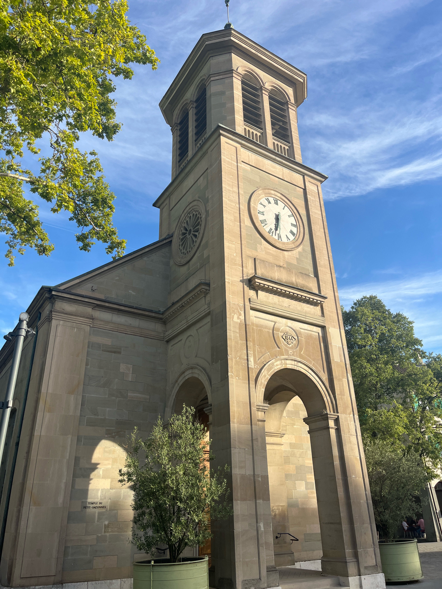 The image shows the exterior of the Temple du Petit-Saconnex, a church located in Geneva, Switzerland. The church features a prominent stone clock tower with Roman numerals on its clock face. To the left of the tower, the church's facade is visible, constructed from large stone blocks. Lush green trees with bright green leaves are present in the foreground and background, casting shadows on the building. The sky is a vibrant blue with wispy white clouds. A small, leafy tree in a green planter sits in front of the church entrance, near a sign that reads "TEMPLE DU PETIT-SACONNEX". In the background, a few people are visible, some seated at a table.