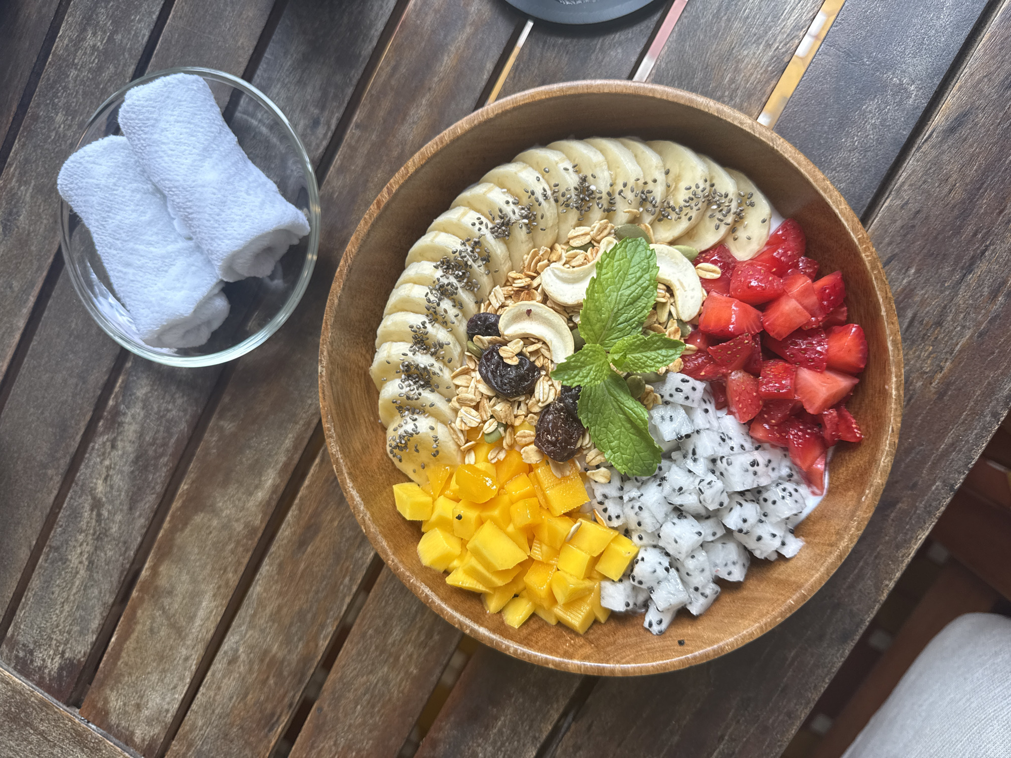 The image depicts a wooden bowl filled with a colorful assortment of fruits, granola, nuts, and a sprig of mint. The fruits include sliced bananas topped with chia seeds, diced strawberries, cubed dragon fruit, and diced mango. Granola, cashews, and raisins are also present in the center of the bowl. The bowl is placed on a wooden table with slats. To the left of the bowl is a small, clear glass bowl containing two tightly rolled white cloths. The scene suggests a breakfast or brunch setting. The location is not specified in the image.