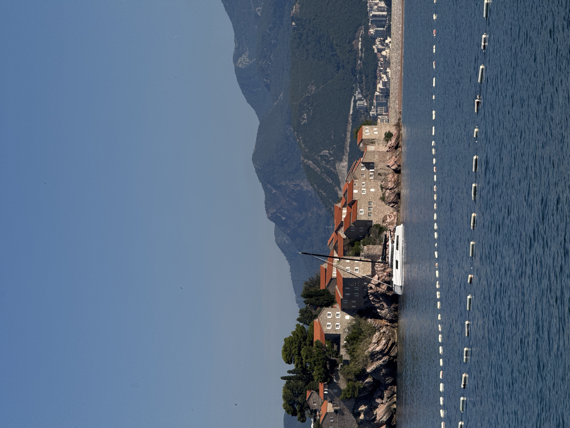 The image depicts the island town of Sveti Stefan in Montenegro, viewed from the water. A modern white sailboat with a dark mast is docked near the island. The island itself is characterized by its historic stone buildings with terracotta roofs, nestled amidst rocky terrain and lush green trees. In the background, a range of densely forested mountains rises, with a distant coastline dotted with buildings visible on the right side of the frame. The sea in the foreground is a deep blue, with a line of white buoys marking a boundary in the water. The sky is clear and a vibrant blue, with a few faint dark specks that may be birds in flight. The overall scene is one of a picturesque coastal landscape on a sunny day.
