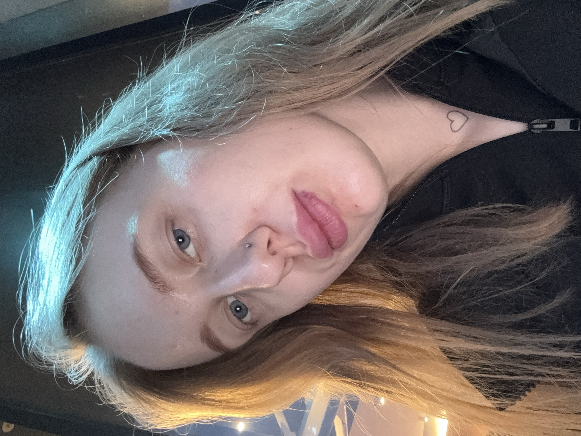 A close-up, rotated portrait of a fair-skinned woman with blonde and brown hair and blue eyes. She has a nose piercing. A small heart tattoo is visible on her neck, near the zipper of a black garment she is wearing. The environment is indistinct but appears to be indoors, with some ambient lighting. There is no indication of Ol'ginskaya, Russia in this image.