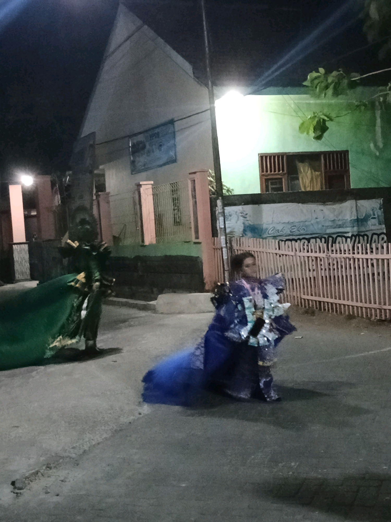 This image depicts an outdoor scene at night, likely in Keplaksari, Indonesia. Two individuals are visible, seemingly participating in a cultural performance or parade.
On the left, a person is dressed in an elaborate green costume with golden accents, holding a large green fabric that drapes to the ground. The costume appears to have shoulder armor or embellishments.
To the right, another individual is adorned in a vibrant blue and silver costume, also with extensive embellishments. This costume features a voluminous skirt and what appears to be a headdress or intricate hairpiece. The person is holding an object that reflects light, possibly a collection of small, metallic items or a decorative accessory.
The environment includes a paved pathway or road, a wooden fence with vertical slats, and a building with a green facade. A banner is visible behind the fence, with text that is partially obscured but includes the words "Cak Eko". The lighting suggests artificial illumination from streetlights or nearby structures, casting shadows on the ground. The overall atmosphere is festive and dynamic, indicative of a public event.