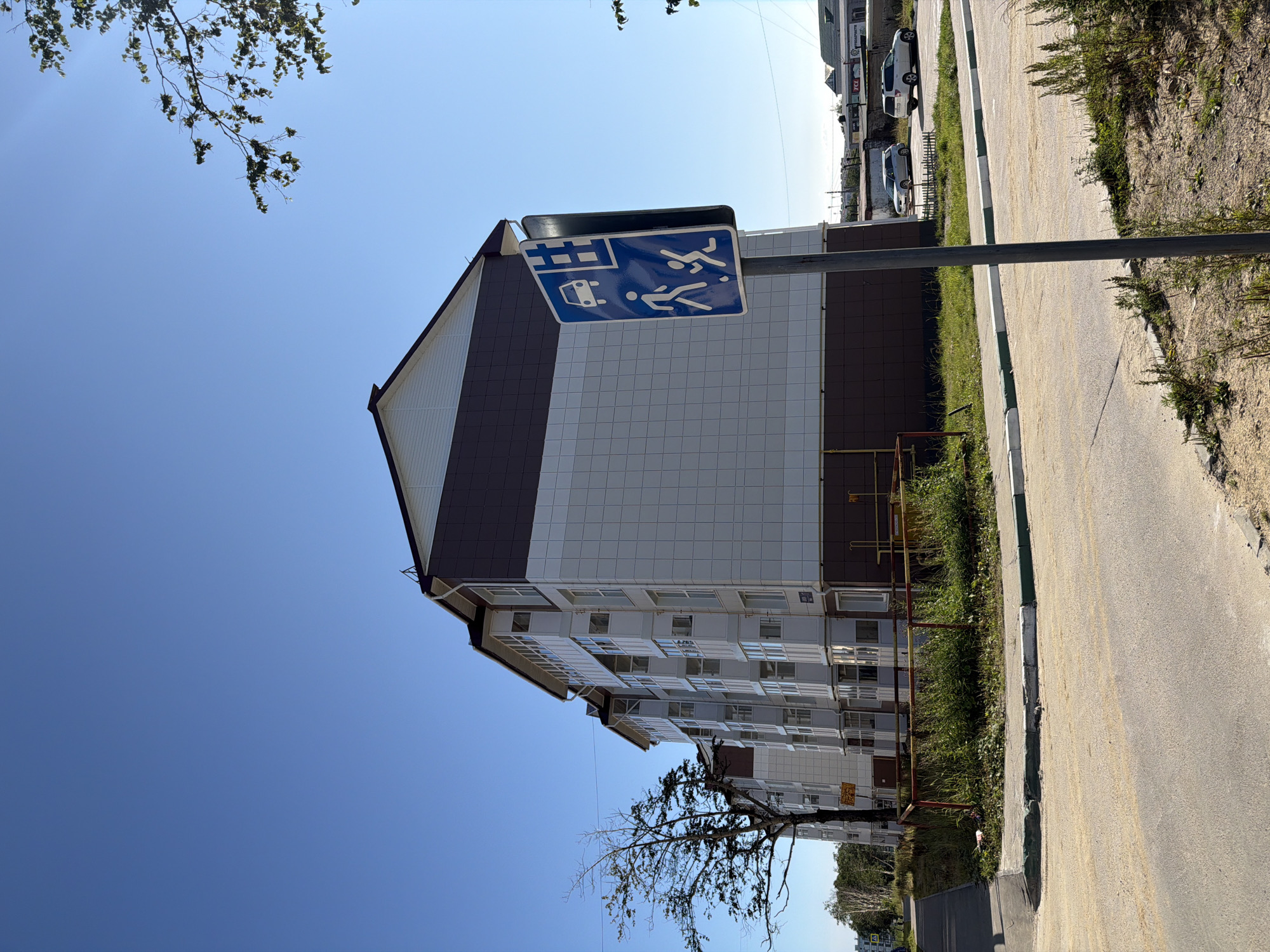 In Okha, Russia, on a clear, sunny day, a modern apartment building with white and dark brown siding stands tall. In front of the building, a traffic sign displaying a car and pedestrian symbols is mounted on a metal pole. To the left of the building, a bare tree with thin branches is visible. A sidewalk with a green and white striped curb runs along the front of the building, and a paved road is in the foreground. Two white cars are parked in the background, near other buildings.