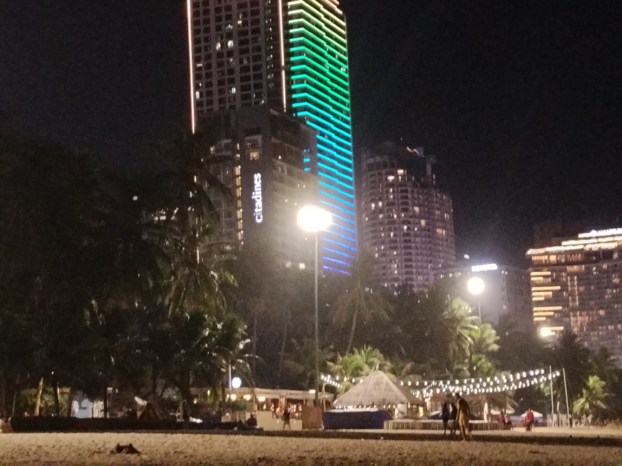 This image depicts a nighttime scene at a beach in Nha Trang, Vietnam, likely on Tran Phu Beach. In the background, several tall, illuminated buildings dominate the skyline. The most prominent is a skyscraper with a facade lit in a gradient of green and blue, displaying horizontal light bands. To its left, another large building shows numerous lit windows. Further to the right, a building with a similar illuminated facade of horizontal bars is visible, alongside other high-rise structures with glowing windows. In the foreground and mid-ground, palm trees are scattered across the scene. Along the beach, there are several structures, including what appears to be a thatched-roof hut or pavilion decorated with strings of small lights. People are visible on the beach, some walking, and others gathered near the structures. The beach itself is sandy and appears to be relatively flat, with some areas of debris or vegetation at the edge of the sand. The overall atmosphere is that of a lively, illuminated urban beachfront at night.