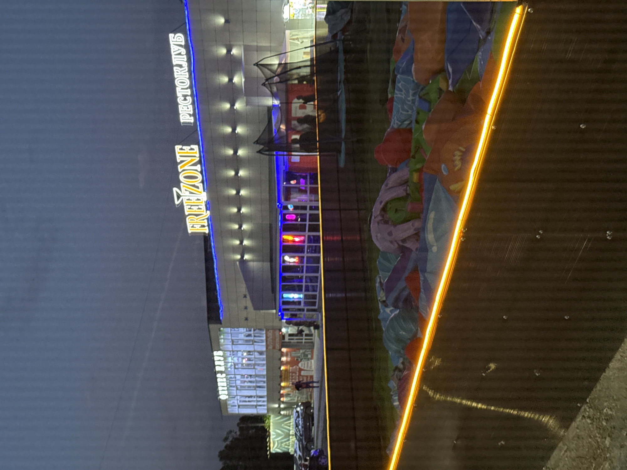 The image captures an outdoor scene at dusk, likely in Mariupol, Ukraine, given the context. The primary subject is a modern building illuminated by vibrant neon lights and floodlights. On the left side of the building, prominent text reads "FREE ZONE" in large, glowing letters, with "PEKTOPLYB" (likely a transliteration or partial word) above it. The building's facade features many windows, some of which reveal illuminated interiors.
In front of the building, a large inflatable play area is visible, filled with colorful structures. People are present, some appearing to be inside the building's illuminated sections, while others are near the entrance or in the vicinity of the inflatable structures. A trampoline with a safety net is also set up. Cars are parked in front of the building, indicating it is a commercial or entertainment venue.
The lighting suggests it is either early morning or evening, with the artificial lights of the building being the dominant illumination source against the darkening sky. The weather appears calm. The overall atmosphere is that of an active recreational space, likely a family entertainment center or amusement park. The presence of multiple neon signs and bright lighting creates a lively and inviting ambiance. The text "FREE ZONE" suggests a place of leisure and activity.