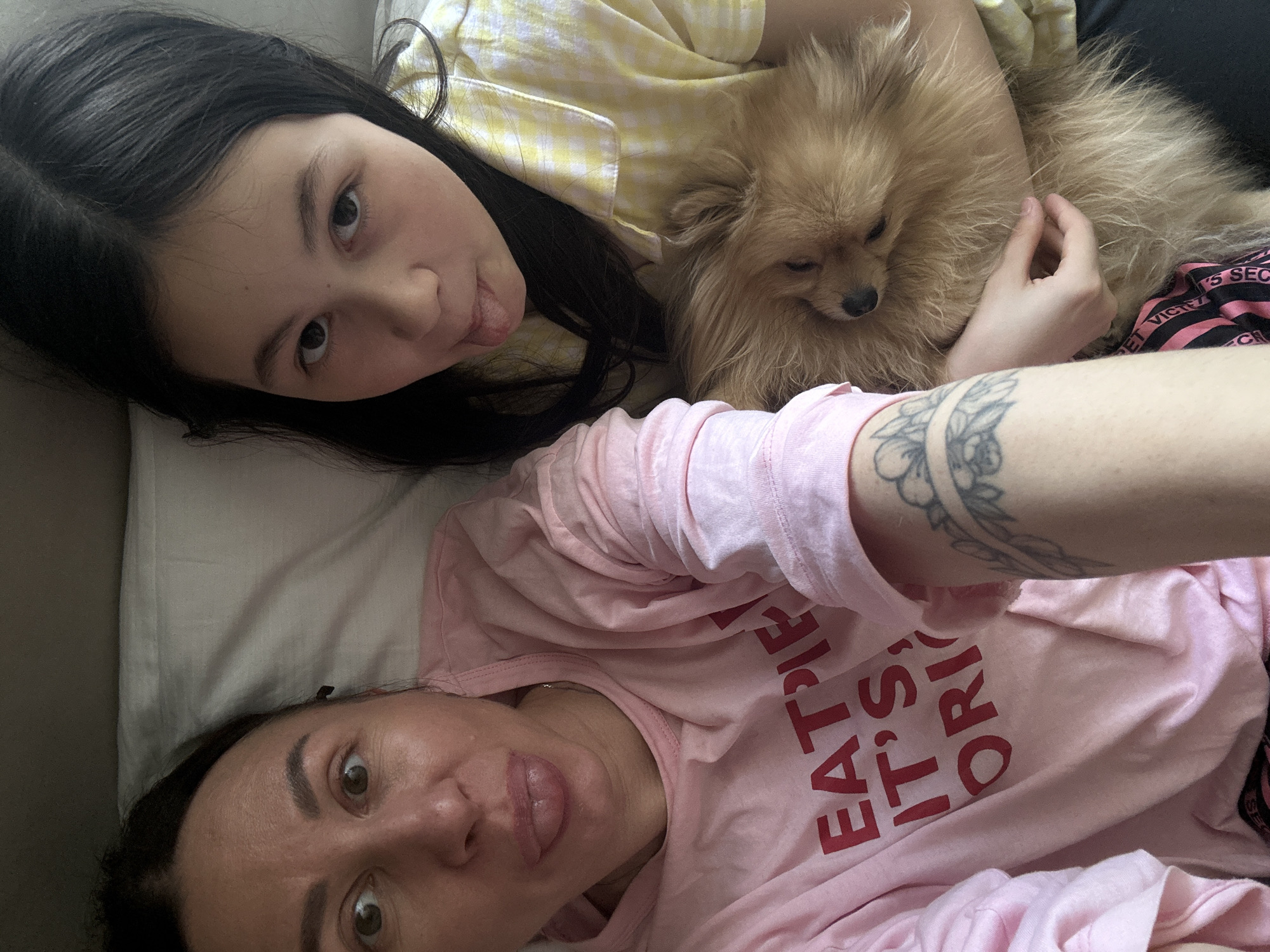 A selfie shows a woman, a girl, and a dog lying on a surface, possibly a bed or couch. The woman, positioned at the bottom of the frame, has dark hair and is wearing a pink shirt with red lettering that reads "EAT PIE, IT'S ORIG". She is sticking out her tongue. A tattoo is visible on her right arm. Above her, a girl with long dark hair is also sticking out her tongue. She is wearing a yellow and white checkered shirt. Between them lies a small, fluffy, light brown dog. The background appears to be a neutral gray, suggesting an interior setting. The location is not evident from the provided image. There is no evidence of the scene being in the city of Votkinsk, Russia.