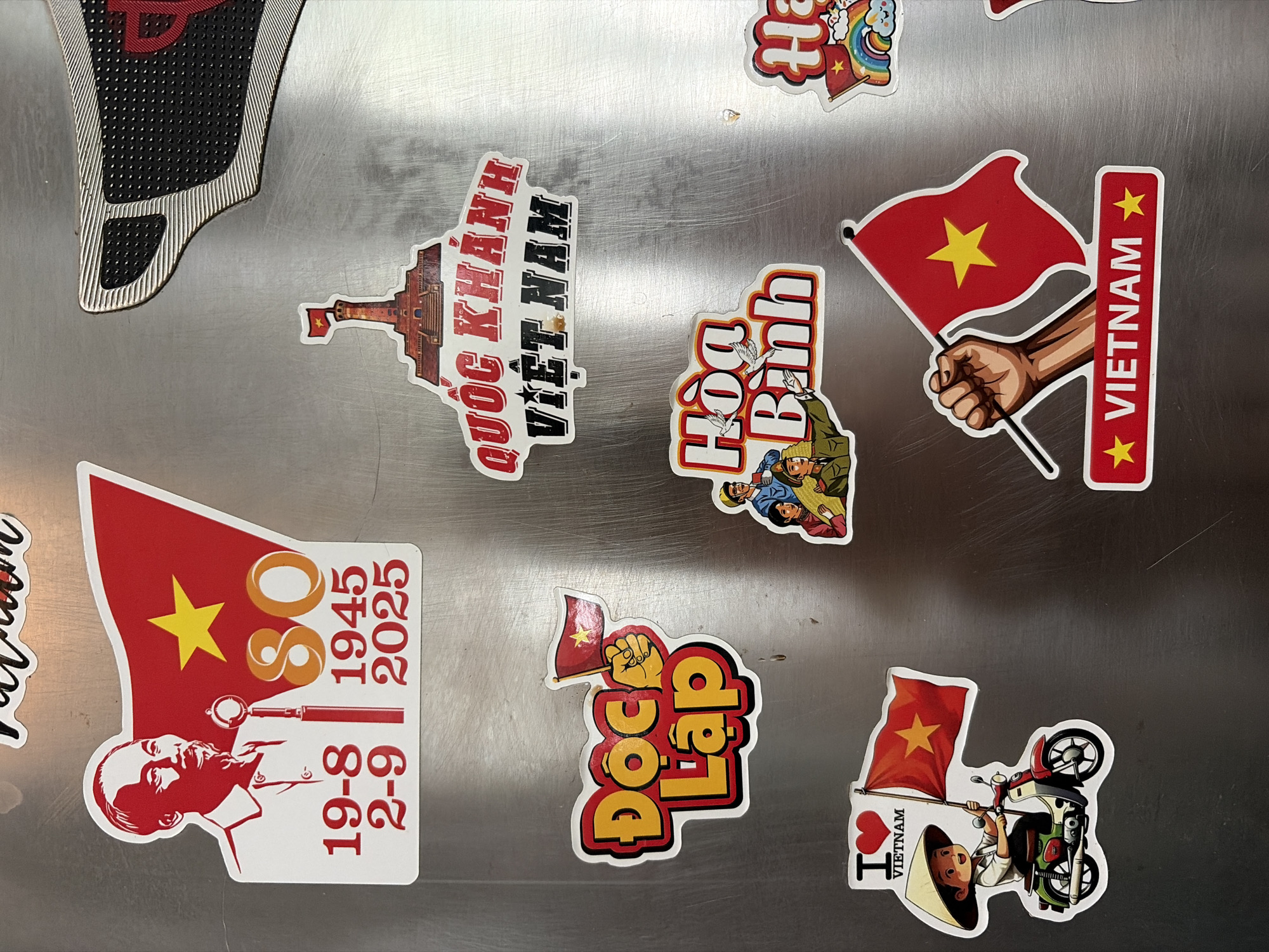 The image shows a collection of stickers on a metallic surface, likely a refrigerator. The stickers feature Vietnamese national symbols and themes, including a portrait of Ho Chi Minh, the Vietnamese flag, and slogans in Vietnamese. One sticker depicts a person riding a scooter with an "I heart Vietnam" message. Another shows a hand holding the Vietnamese flag with "VIETNAM" written below. The overall theme appears to be national pride and celebration. The location in Đà Nẵng is not determinable from the image.
