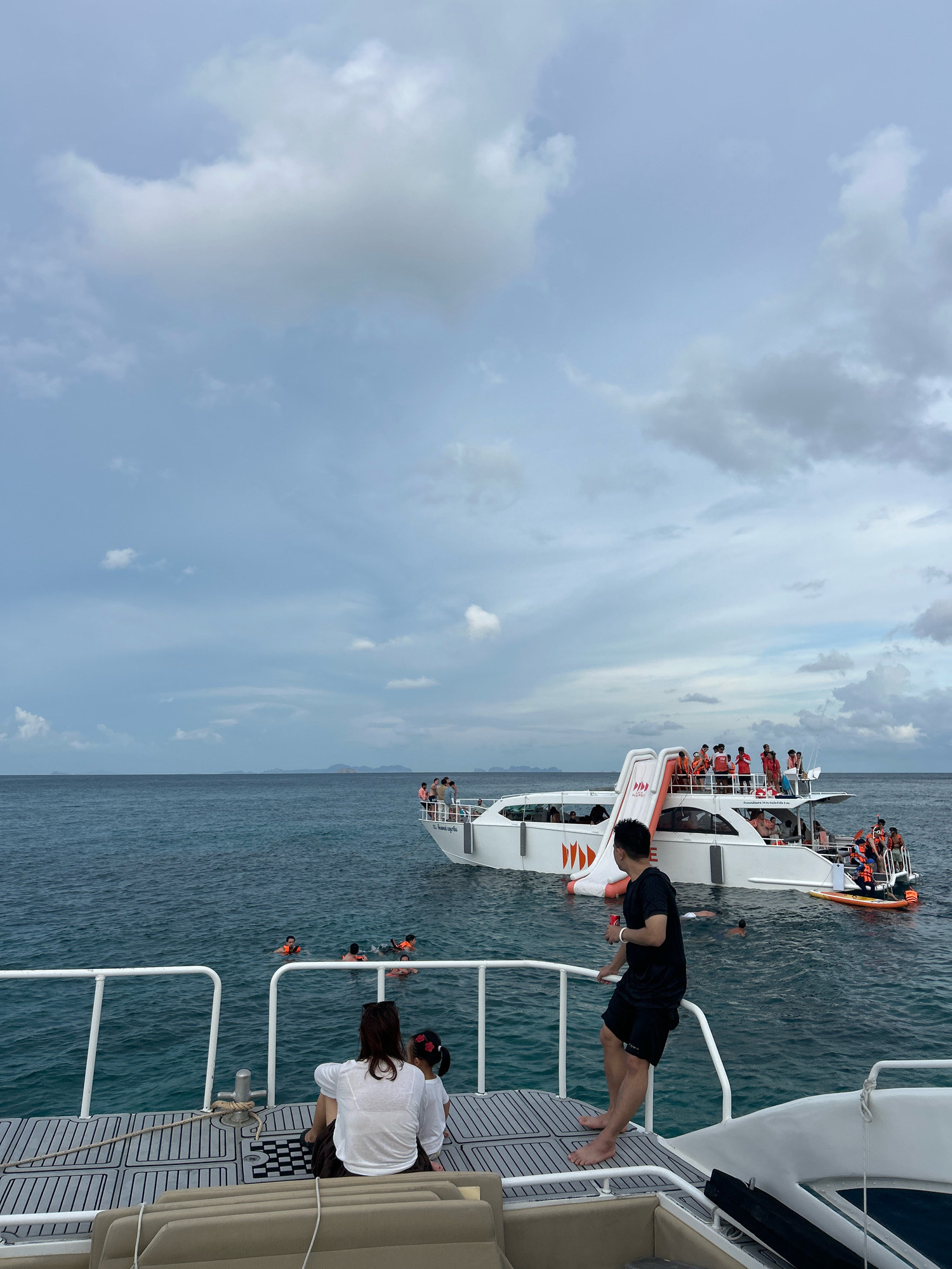 In Rawai, Thailand, a group of tourists are enjoying a day at sea. Two people are seated on the deck of a boat in the foreground, facing away from the camera. Behind them, several individuals are swimming in the ocean wearing life vests. A larger white boat with an orange slide is anchored in the middle ground, with many people visible on its decks, some wearing life vests. The sky is overcast with dramatic clouds, and the sea is a deep blue. In the distance, several islands can be seen on the horizon.