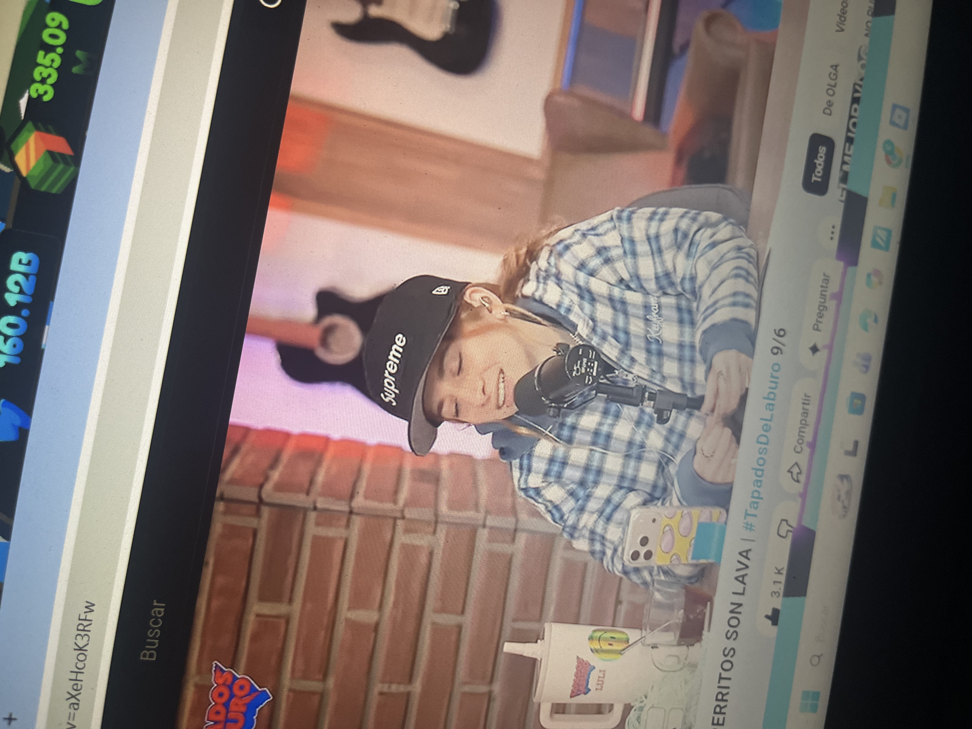 The image shows a woman wearing a plaid shirt and a black baseball cap that says "Supreme". She is sitting in front of a brick wall and holding a microphone. There are two guitars visible in the background. The woman appears to be on a video call or streaming. There is no information in the image that places the scene in Santa Ana, Venezuela.
