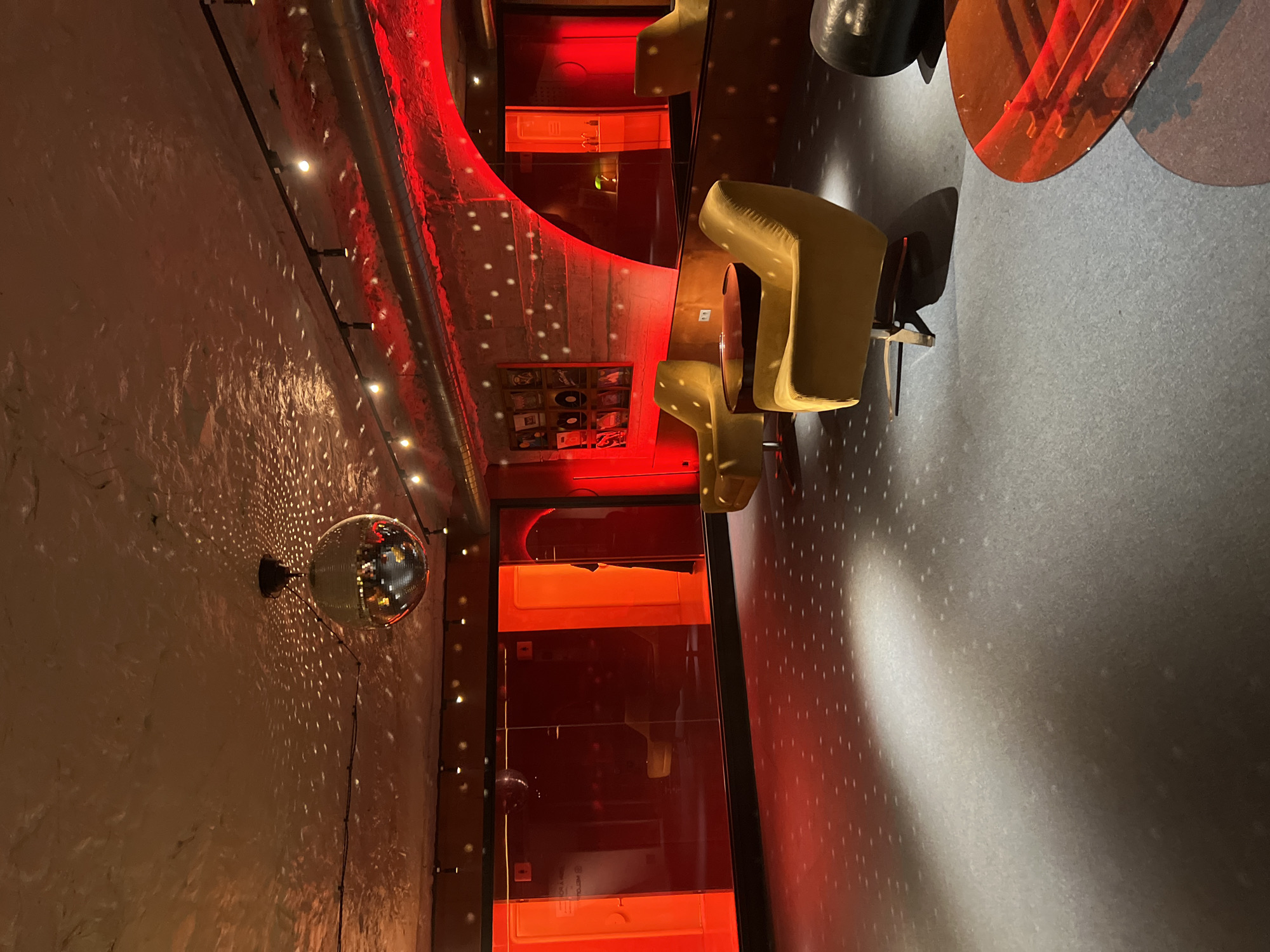 The image depicts an interior space with a retro or vintage aesthetic, possibly a bar or lounge, in Yerevan, Armenia. A disco ball hangs from the textured wall, casting points of light across the space. The lighting is predominantly red, illuminating walls, furniture, and reflective surfaces. Several mustard-yellow chairs are visible, arranged around a central area. A shelving unit displaying what appear to be records or similar items is seen on the wall. The floor is a neutral gray, speckled with light. No people are visible.