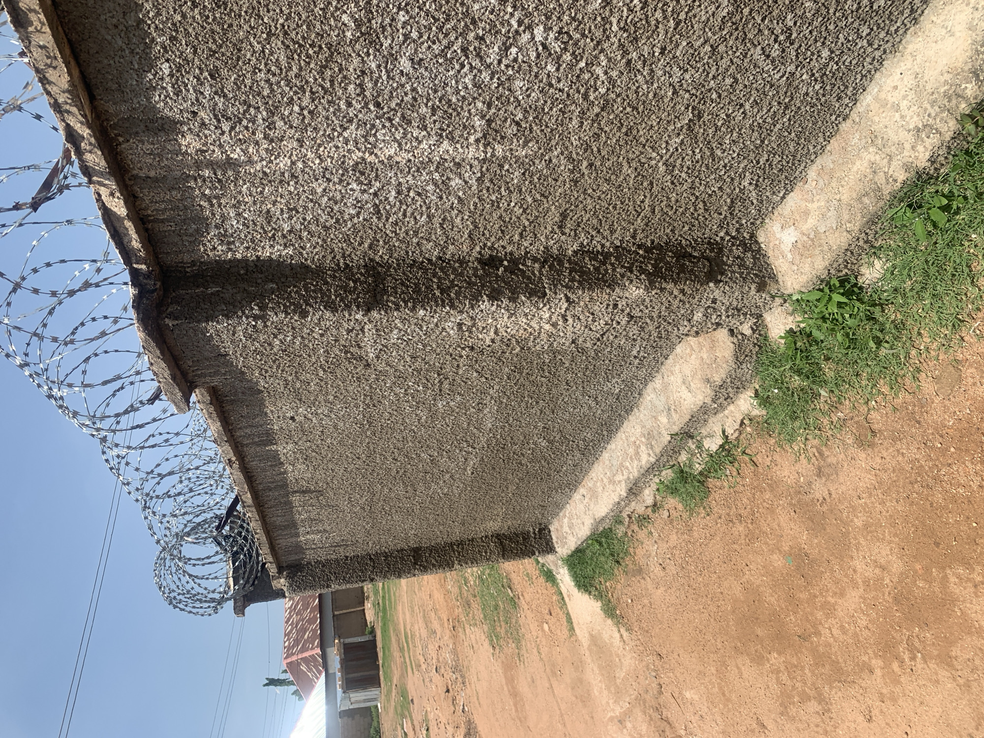 The image shows a rough, concrete wall topped with coiled barbed wire. The wall is textured and appears to be part of a perimeter fence. In the background, there are buildings with corrugated metal roofs and a dirt road. The sky is clear and blue. The overall scene suggests an outdoor, possibly residential or commercial, area in Jauro Yinu, Nigeria. The focus is on the wall and security measures, with the surrounding environment providing context.