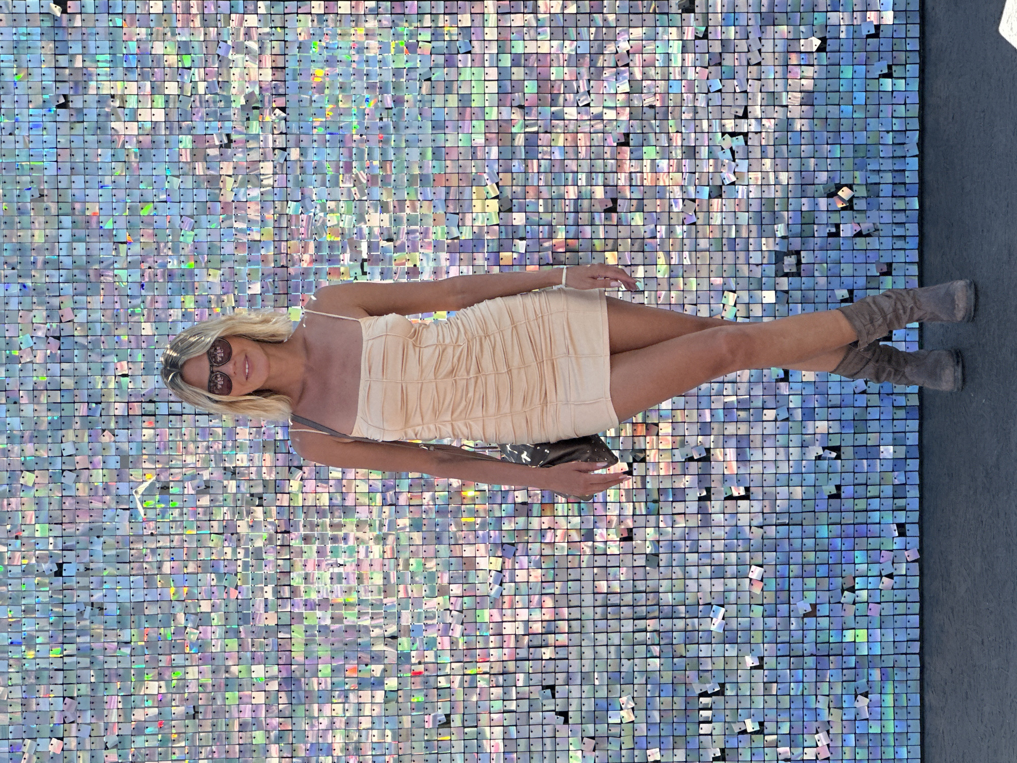 A woman stands facing forward in front of a wall covered in shimmering, square sequins. She has blonde hair and is wearing sunglasses, a pale peach-colored ruched mini-dress, and grey slouch boots. A dark purse with studs is slung over her left shoulder. The background is a wall of iridescent sequins that reflect light and colors. The floor in front of the wall is dark and textured. There is no indication of the city of Moscow, Russia in the image.