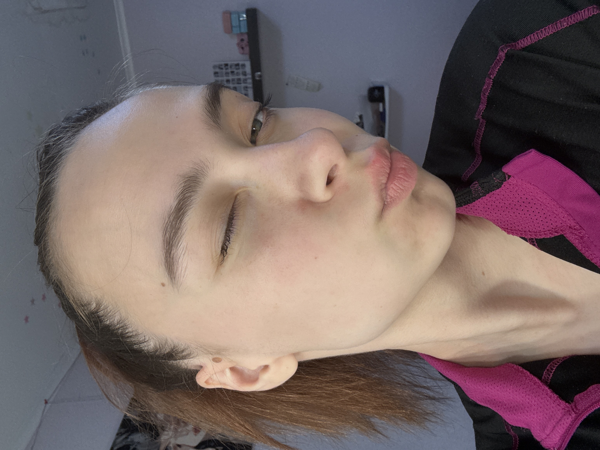 A close-up, tilted portrait shot of a young woman with fair skin, light brown hair, and partially closed eyes. She is wearing a black and magenta athletic top. Her expression is neutral, with slightly parted lips. The background is blurred but appears to be an indoor setting, possibly a bedroom, with faint outlines of a shelf or wall decorations. There is no information to support the image was taken in Прикамский, Russia.