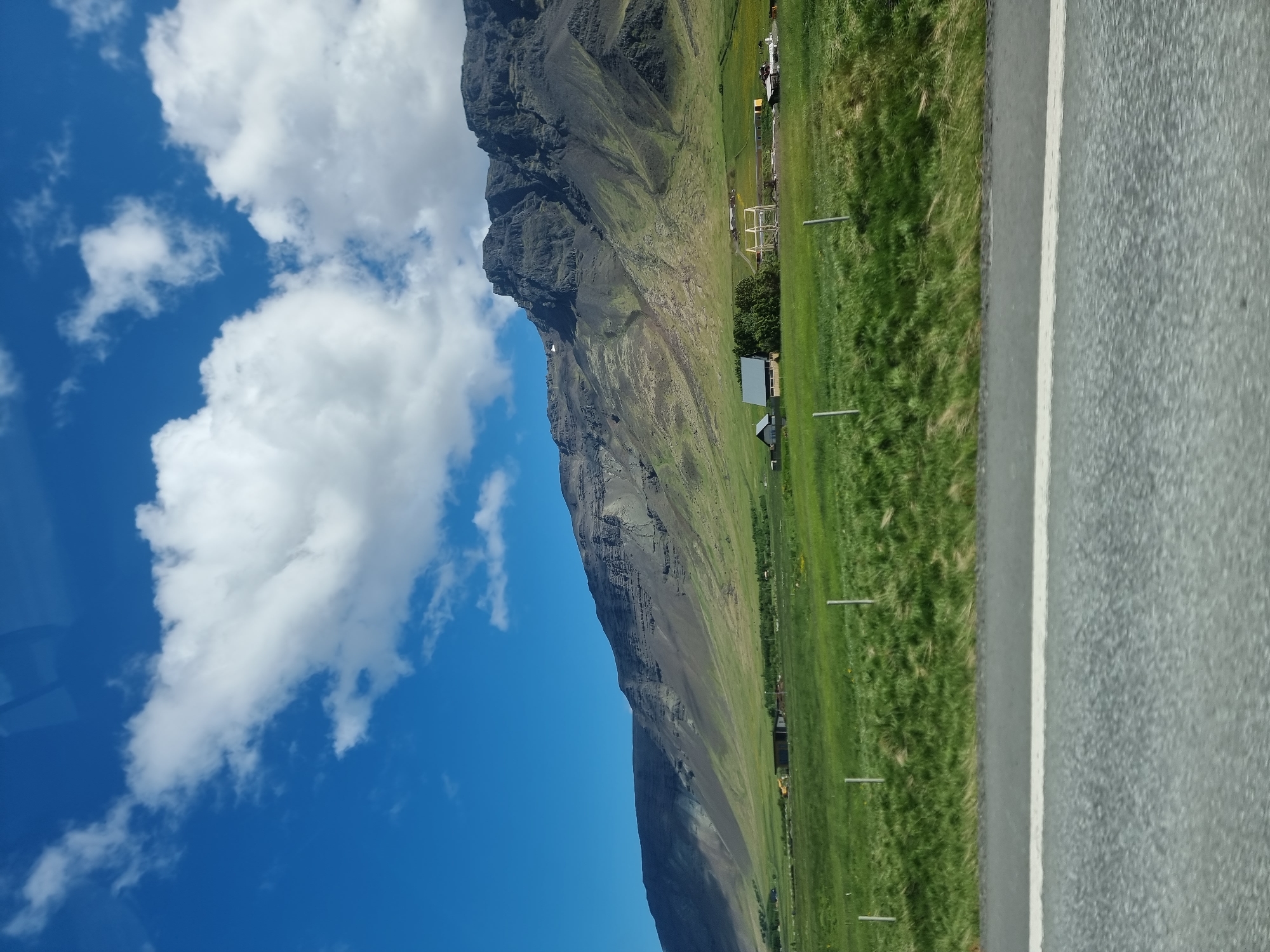 The image depicts a vast, green grassy plain leading up to a rugged, dark grey mountain range under a bright blue sky with scattered white clouds. A paved road with a white stripe runs across the foreground. In the middle ground, there are several low-rise buildings, including a house with a grey roof and a long, shed-like structure. A few trees are visible near the buildings. The scene appears to be a rural landscape, possibly an agricultural area or a residential area on the outskirts of a town or city. There are no people or animals visible in the image. The lighting suggests it is daytime, likely sunny. This image is not located in the city of Reykjavik, Iceland, as it depicts a remote natural landscape with mountains, which are characteristic of areas outside of Reykjavik's urban center.