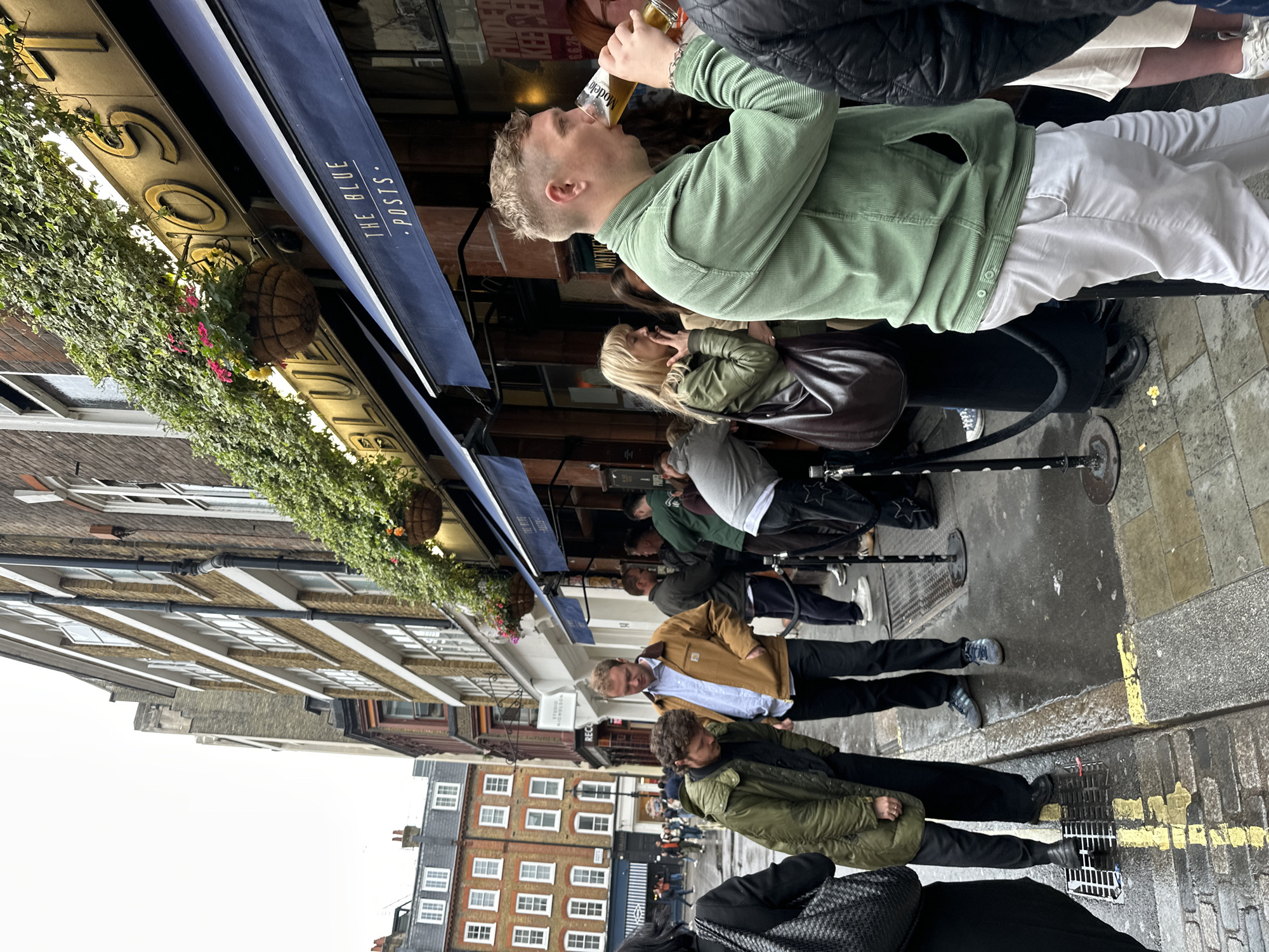 This image captures a street scene in London, United Kingdom, specifically near The Blue Posts pub on Rupert Street in the Soho district, evidenced by the pub's signage and the "STUDIO NICHOLSON" sign visible in the background.
The scene depicts a moderately busy urban street, with multi-story brick buildings lining both sides under an overcast sky. The pavement appears wet in areas, indicating recent rain. On the right, The Blue Posts pub is prominent, featuring a dark blue awning with "THE BLUE POSTS" written in white lettering. Above the awning, a gilded sign with raised lettering spelling out "BLUE POSTS" is partially covered by lush green ivy and adorned with hanging baskets containing vibrant red and yellow flowers.
In the immediate foreground to the right, a man with short, light-colored hair, wearing a light green corduroy jacket and white trousers, is actively drinking from a glass of beer. To his left, a woman with blonde hair, wearing a green jacket and carrying a dark brown shoulder bag, is smoking a cigarette. Further left, two men stand near the curb; one with dark, curly hair wears an olive-green hooded jacket and black trousers, looking downwards. The other, with light brown hair, wears a tan jacket over a light blue collared shirt and black trousers, also looking towards the ground.
A series of black bollards with dark rope form a queue barrier outside the pub. The street surface is a mix of wet cobblestones and concrete, marked with yellow lines near the curb, and a metal drain grate is visible in the foreground. Several other individuals are visible in the mid-ground and background, standing, walking, and congregating along the street. Architectural details include typical London brickwork, numerous sash windows, and downpipes on the building facades.