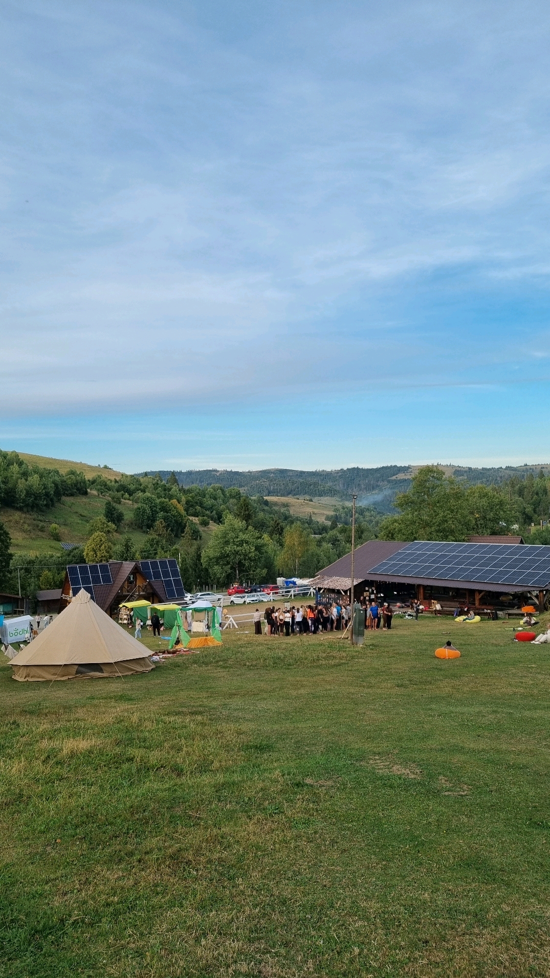 The image depicts an outdoor gathering at what appears to be a rural campsite or event ground in Izky, Ukraine. A large bell tent is set up in the foreground on a grassy field. Several smaller tents, some with green canopies, are situated behind the bell tent. A group of people are standing and interacting in the middle ground, near a building with a sloped roof covered in solar panels. This building appears to be a communal area or restaurant. Beyond this building, a line of parked cars is visible. In the background, rolling hills covered in dense green trees stretch towards the horizon under a partly cloudy blue sky. The overall scene suggests a daytime event, possibly a festival or retreat, taking place in a natural, mountainous environment.