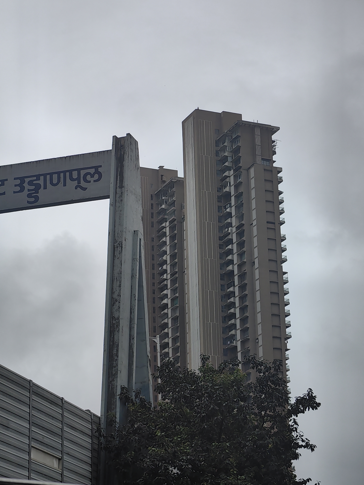 The image shows a street sign in Marathi which reads "उड्डाणपूल" (Uddanpul), meaning "flyover" in English. The sign is attached to a grey, weathered concrete pillar. In the background, two tall, modern apartment buildings dominate the skyline, their beige facades covered in rows of balconies. The sky is overcast and grey. In the foreground, a dark green tree with dense foliage partially obscures the lower part of the scene. To the left of the pillar, a corrugated metal barrier is visible. The scene appears to be a street view in Mumbai, India. There are no people or animals visible in the image.