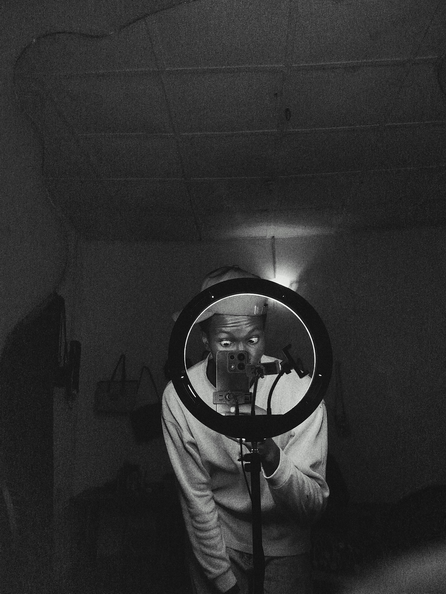 A black and white indoor shot shows a person taking a mirror selfie. The person is wearing a hat and sweater and is holding a ring light with a smartphone attached to it. The person has a surprised expression on their face with their eyes wide open, and looking to the side. The room has a ceiling with what looks like a water stain. There are items on the wall, including a handbag and what looks like a painting or print. It is unknown where the image was taken or if it is in Unknown, Unknown.