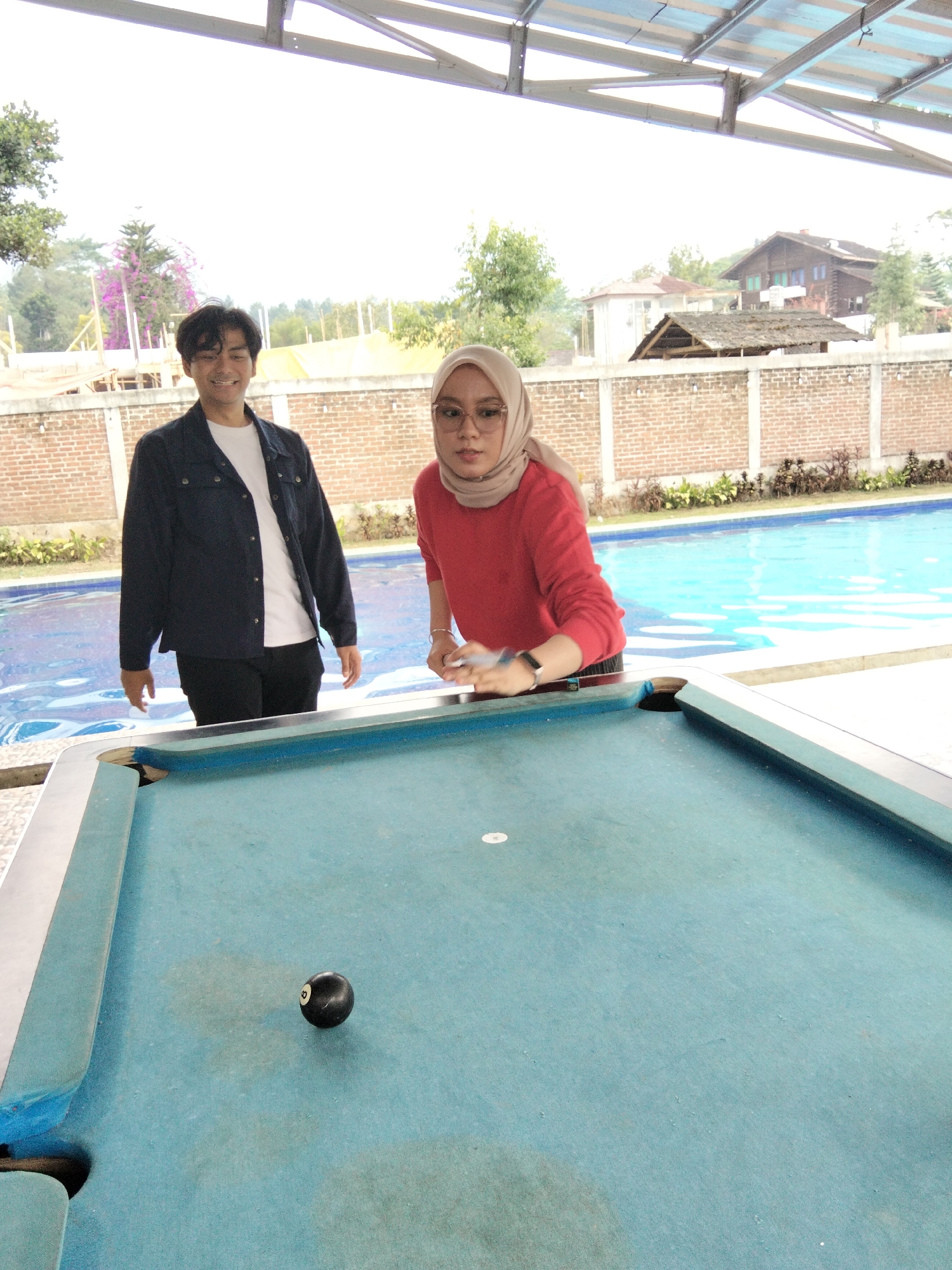 In Cisarua, Indonesia, two individuals are by a swimming pool, with a pool table in the foreground. A young man with dark hair, wearing a dark jacket over a white t-shirt and black pants, stands to the left, smiling at the camera. To his right, a young woman in a beige hijab and a red long-sleeved shirt, wearing glasses, is poised to strike a billiard ball on the pool table. The pool table is light blue with a few visible stains. A single black billiard ball with the number '8' is positioned on the table. Behind them, a bright blue swimming pool stretches out, bordered by a brick wall. In the background, houses and trees are visible, indicating a residential area or resort. The lighting suggests an overcast day.