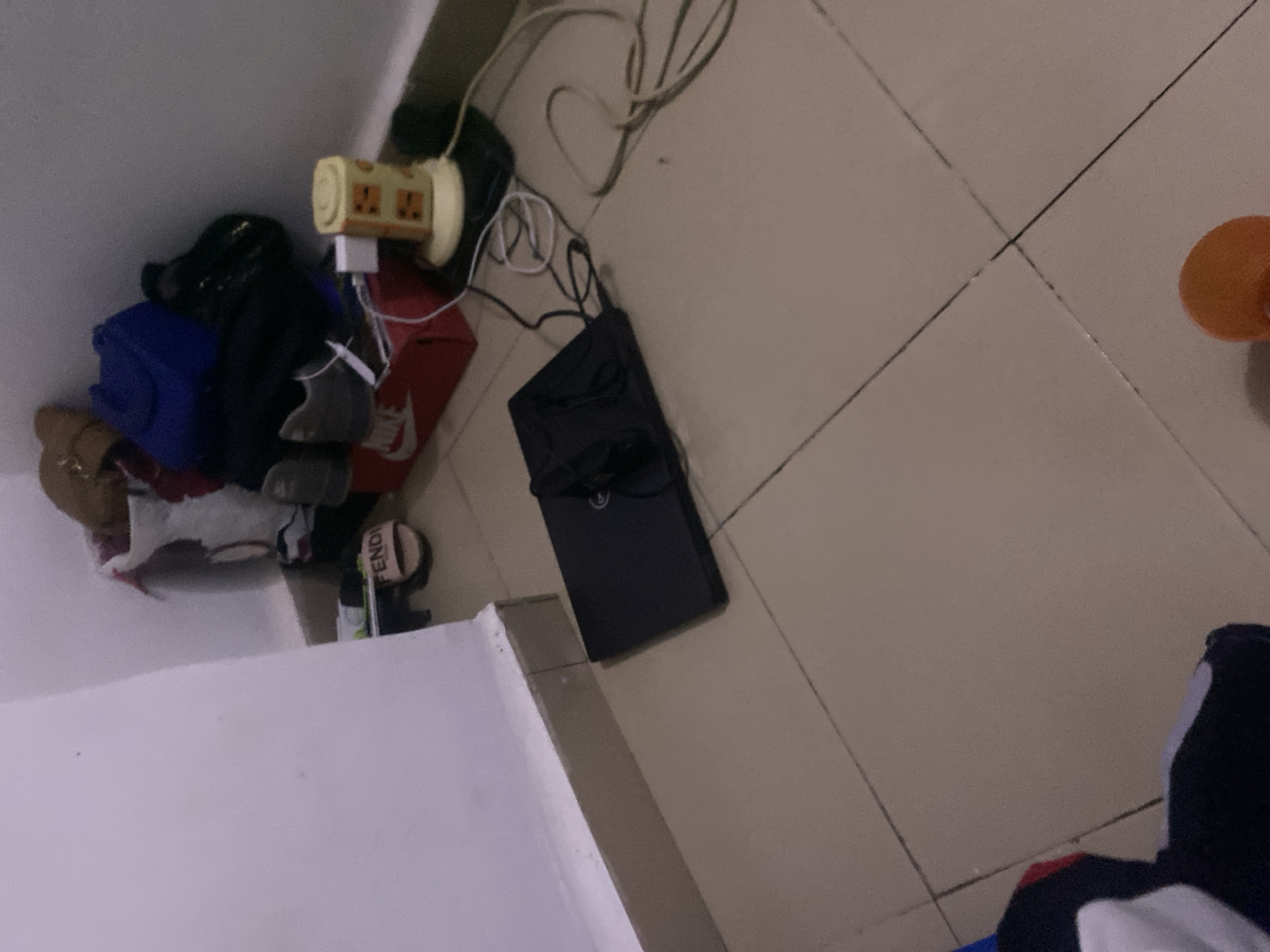 The image shows a cluttered corner of a room with tiled flooring. In the center, a black laptop is placed on the floor. To the right of the laptop, a yellow electrical adapter with multiple outlets is plugged into the wall and connected to various cords. Behind the laptop and adapter, there are several items piled against the wall, including a red shoebox, a pair of gray sneakers, a blue bag, and other dark-colored bags and fabric items. A pink Fendi sandal is visible on the floor near the shoebox. The walls are plain white. The scene depicts a disorganized personal space. The location is identified as Howayi, Nigeria.