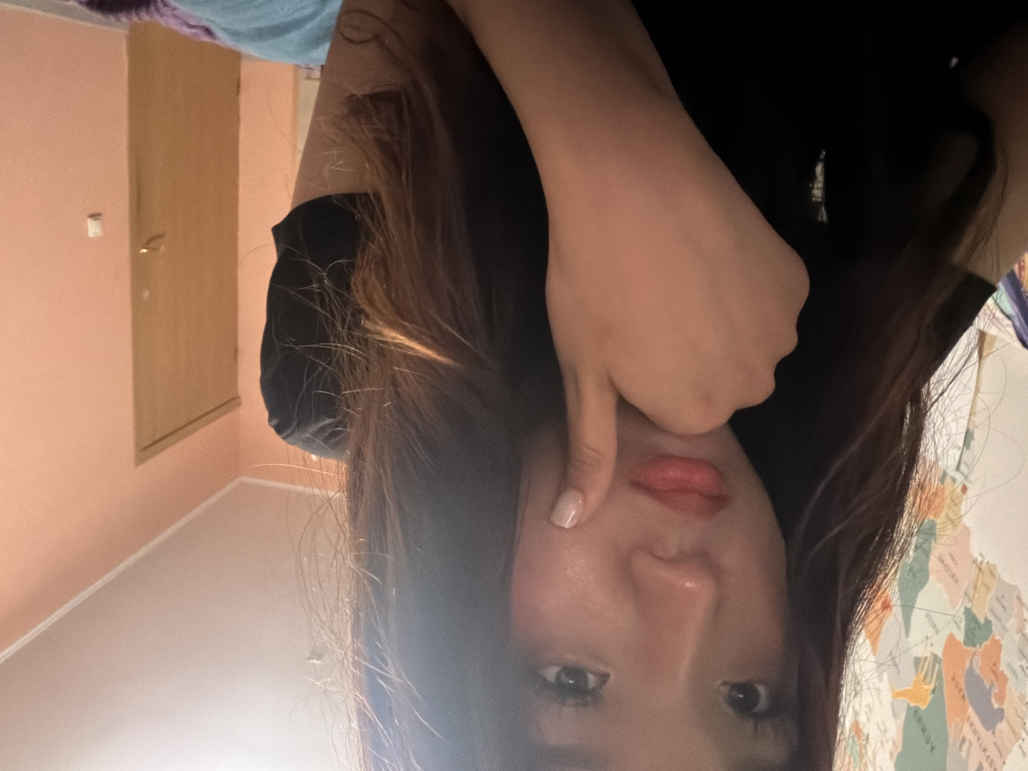 The image shows a young woman in a close-up selfie, with her face and upper body visible. She has dark hair and is wearing a black top. Her right hand is touching her face, with her finger resting on her cheek. She appears to be looking down. The background is slightly out of focus, but it shows a wall with a world map and a peach-colored wall with a door and a light switch. The lighting suggests an indoor setting, possibly a bedroom. There is no indication of the specific location in the city of Unknown, Russia.