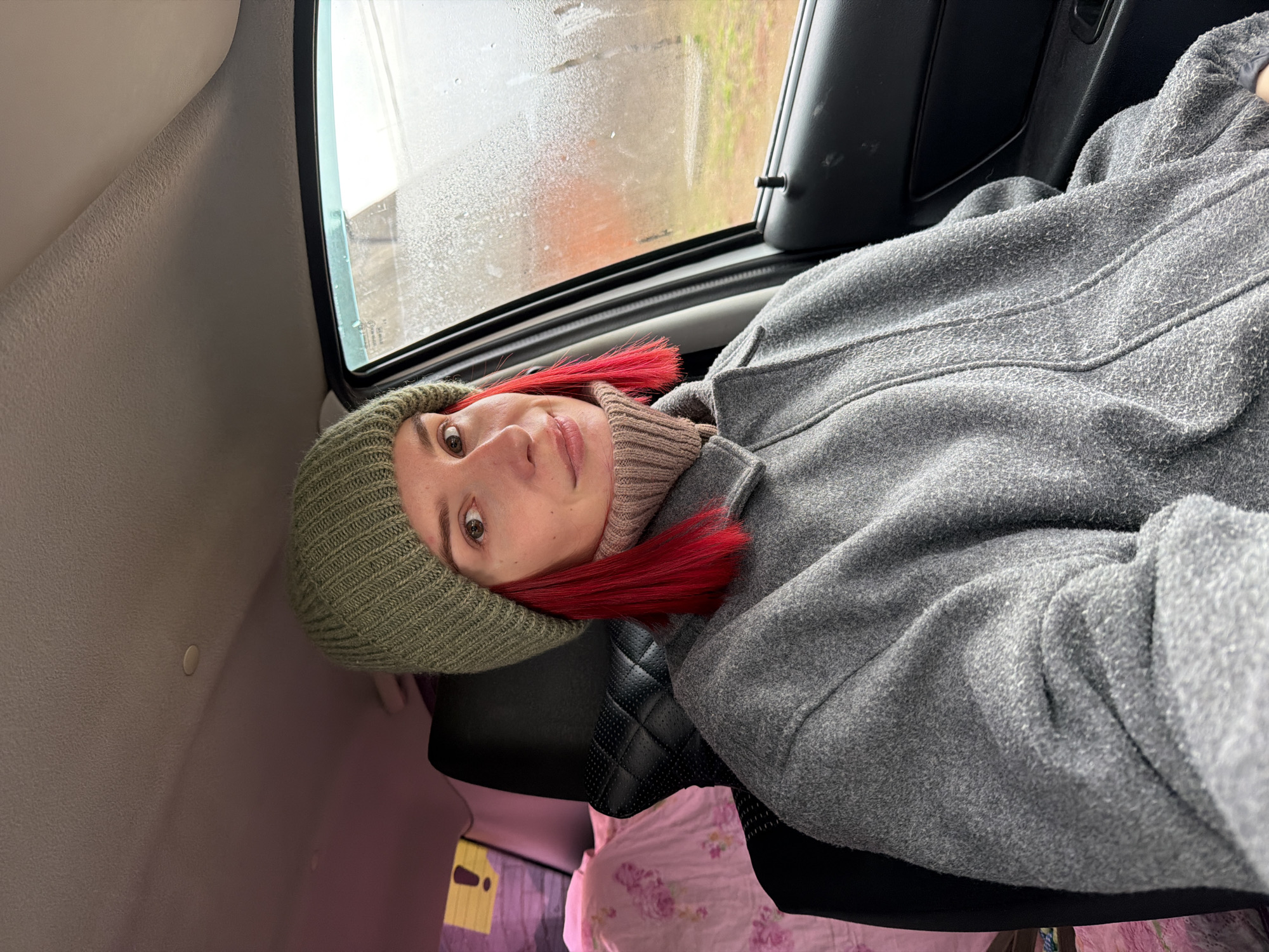 A woman is lying down in the backseat of a car. She is wearing a green knit hat, a beige turtleneck sweater, and a gray wool coat. Her hair is dyed red. The car window is visible, and it appears to be raining outside. The location is Chaikovsky, Russia, based on the presence of Cyrillic lettering and details in the environment.