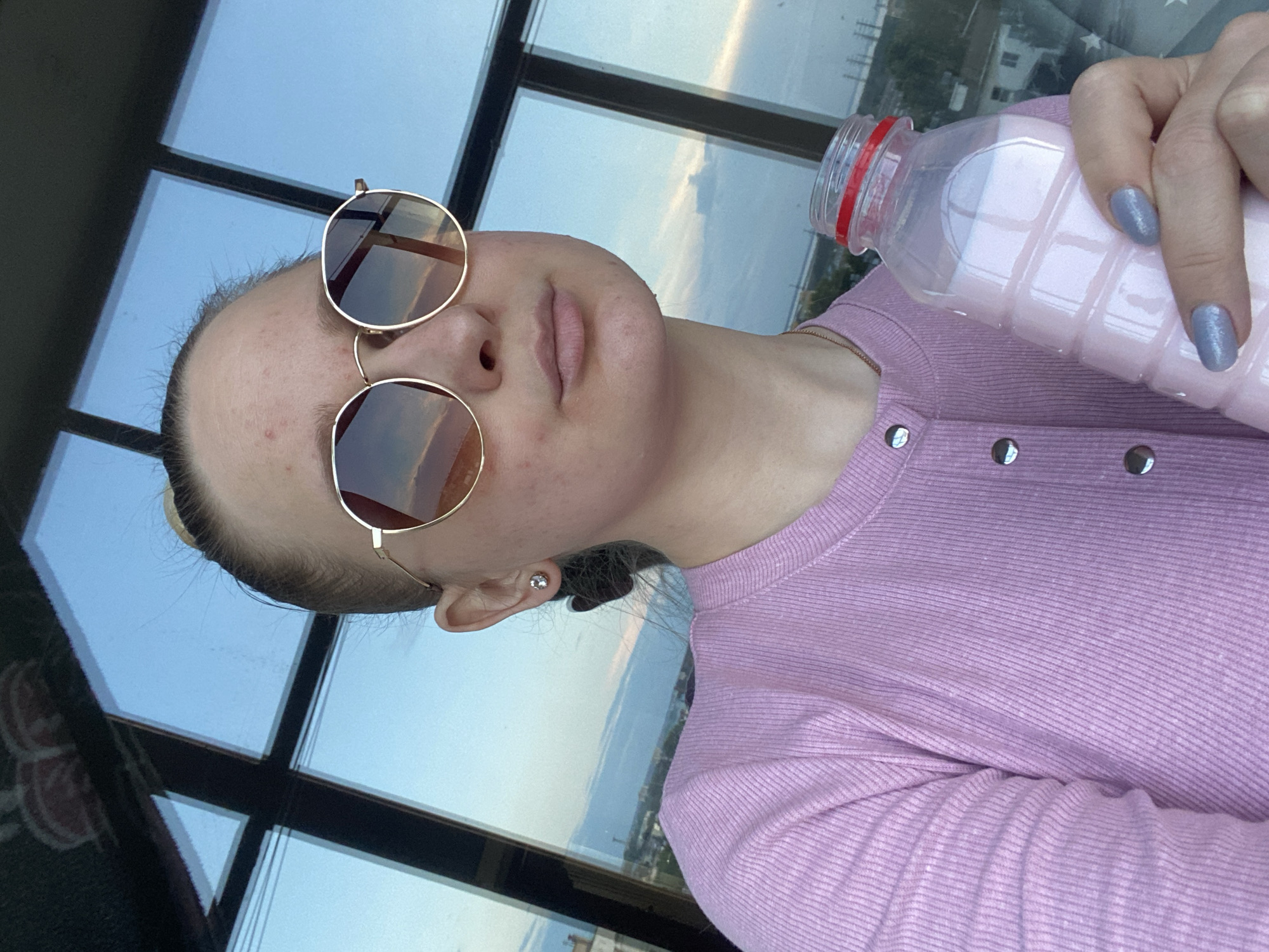 A young woman with blonde hair pulled back into a bun and wearing round sunglasses and a pink ribbed shirt holds a bottle of pink liquid. She is indoors in front of a large window that overlooks a city skyline. The window is framed by dark mullions. The sky outside is blue with scattered clouds, reflecting the sunset hues. Her fingernails are painted a light blue. The image is taken from a low angle, looking up at the woman.