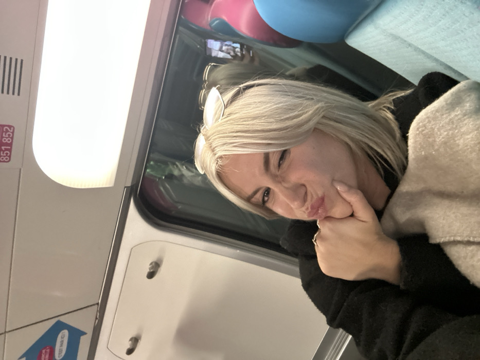 A blonde woman poses with pursed lips and a slight wink, her face resting on her hand. Sunglasses are perched atop her head. She's seated inside what appears to be a train car, judging by the overhead light and interior paneling. A light-colored blanket is draped over her shoulder. Pink and blue objects are visible through the train window behind her.