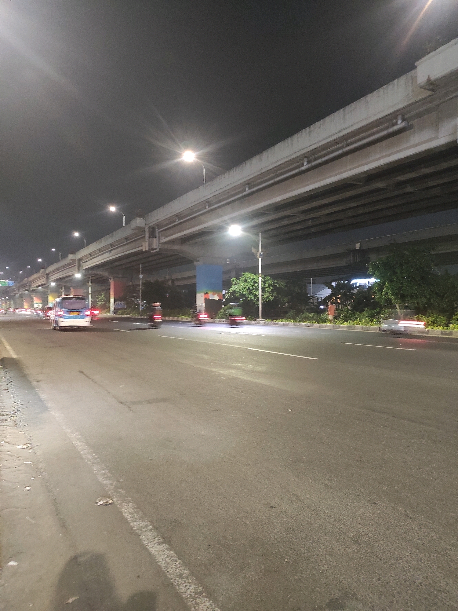 The image depicts a nighttime scene on a multi-lane road in East Jakarta, Indonesia, beneath an elevated highway structure. The road surface is asphalt, with white lane markings visible. Several vehicles are present on the road, including a blue and white van in the left lane, a motorcycle blurred in motion in the right lane, and other vehicles with visible headlights and taillights in the distance. Streetlights illuminate the scene from above. The elevated highway, constructed with concrete beams and supporting columns, casts shadows on the road below. Lush green trees and vegetation line the side of the road. The sky is dark and overcast.