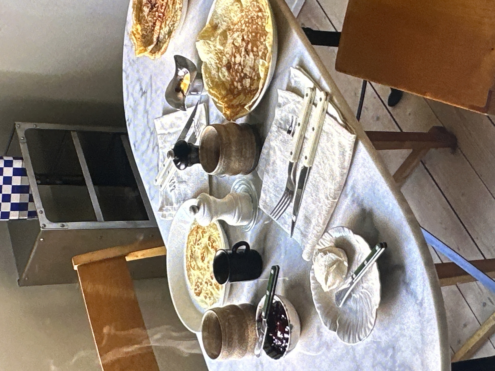 The image depicts a breakfast table setting in an indoor environment. There are plates of crêpes, along with condiments like jam and what appears to be whipped cream. A coffee mug and a sugar or salt shaker are also present. Silverware is arranged on napkins, and there's a pepper grinder visible. The table itself is circular, with a light-colored surface. The background shows elements of furniture, including a wooden chair and a metallic cabinet with a checkered design. There are no visible people or actions taking place. There is no confirmed information about Yerevan, Armenia.