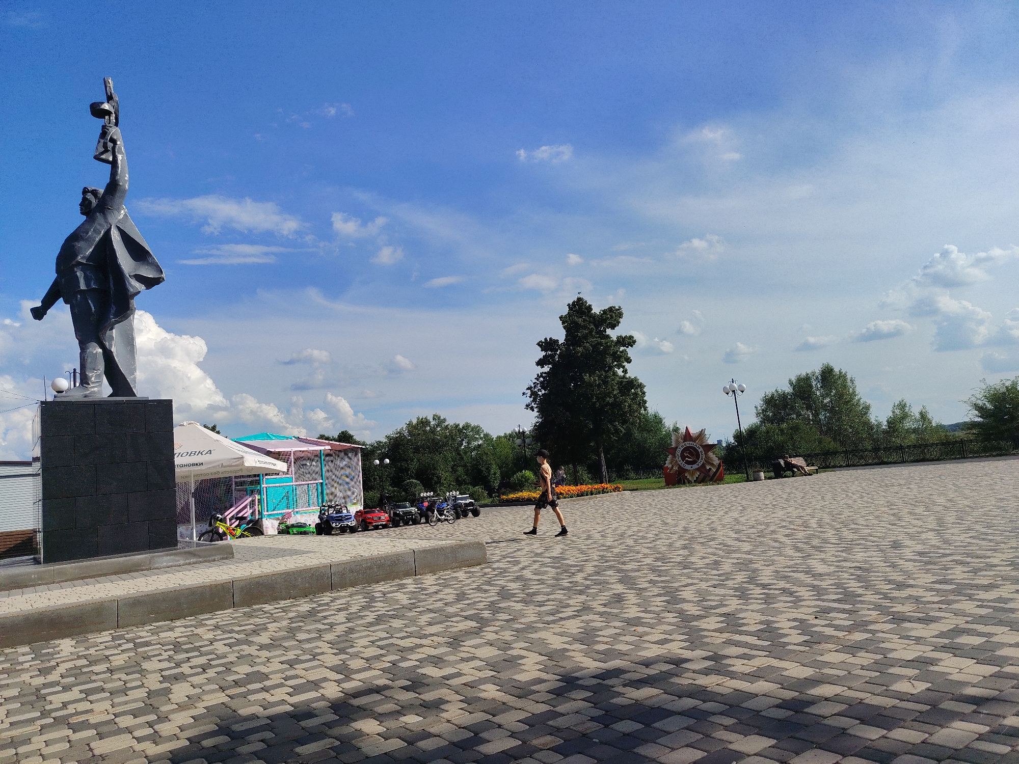 This image depicts a wide, paved plaza in Kemerovo, Russia, featuring a prominent monument on the left. The monument is a dark, angular statue of a figure raising an object aloft, set on a dark grey rectangular base. To the right of the monument, there's a colorful amusement area with a tent, possibly a play area for children, with several small ride-on vehicles like jeeps and ATVs parked in front. A single individual, a male wearing a t-shirt, shorts, and a cap, walks across the plaza away from the monument. In the background, there are trees, a red star-shaped monument with Soviet insignia, a fence, and a bench with a person sitting on it. The sky is blue with scattered white clouds. The plaza is paved with interlocking grey and beige stones, and a concrete curb runs along the edge of the monument's base. The overall scene suggests a public space, likely a park or memorial area, on a bright, sunny day.