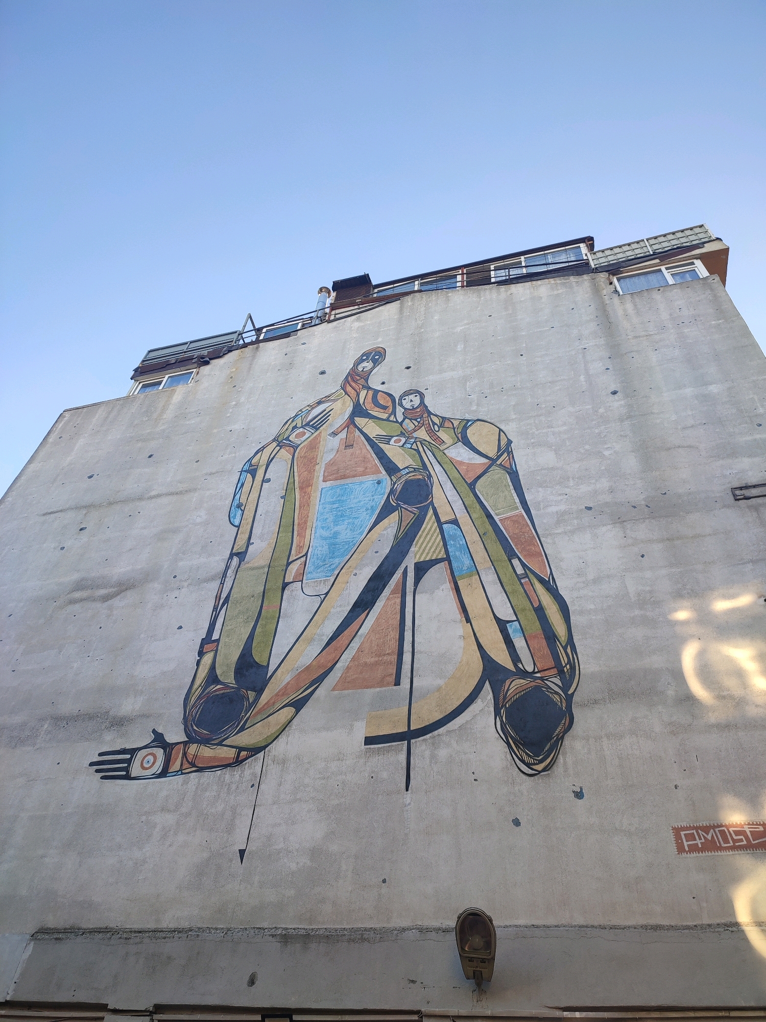 The image shows a large mural painted on the side of a building in Kadıköy, Turkey. The mural depicts an abstract, colorful figure that appears to be composed of geometric shapes and lines. The figure is predominantly yellow, brown, green, and blue, with black outlines. It has a distinctive head with small facial features, and a body that seems to be draped or cloaked. One arm is extended outwards, with a hand depicted with five fingers. The mural is on a rough, concrete wall that shows signs of age and weathering, with scattered dark spots. At the top of the building, there are windows and a rooftop structure with a chimney. A street lamp is attached to the wall near the bottom. In the lower right corner of the mural, the artist's signature "AMOSE" is visible within a rectangular, stamp-like design. The sky above is clear and blue.