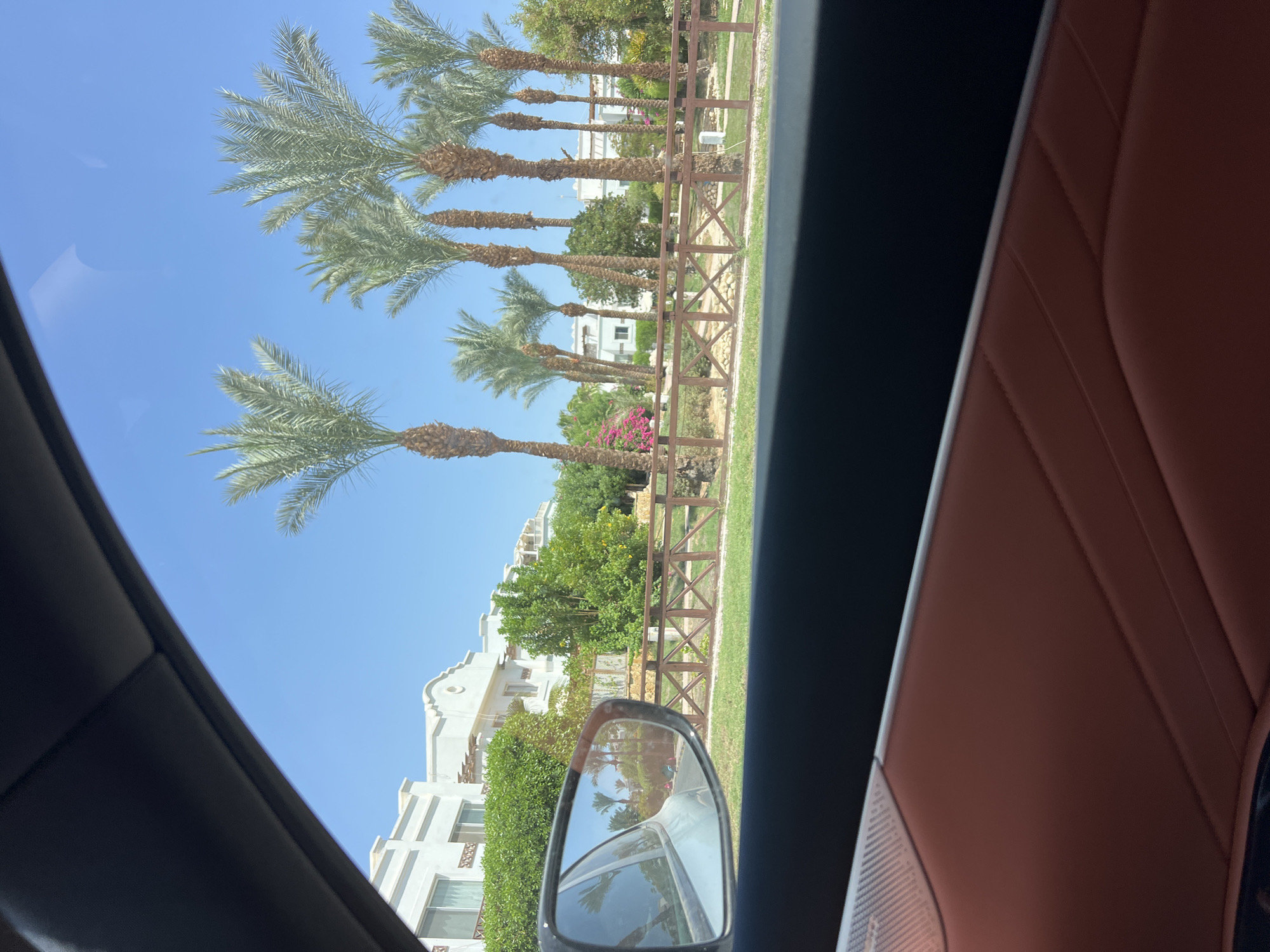 The image captures a scene from inside a car in Sharm El Sheikh, Egypt, looking out the window. In the foreground, the car's interior is visible, featuring a brown leather door panel with stitching. The side-view mirror reflects palm trees, a wooden fence, and a building. Beyond the mirror, a row of tall palm trees stands against a clear blue sky. Behind the palm trees, a white building with an arched detail and several windows is visible, with lush green foliage and some pink flowers in front of it. A wooden fence runs horizontally across the middle ground, separating the road from the landscaped area. The overall impression is of a sunny day in a resort-like setting.