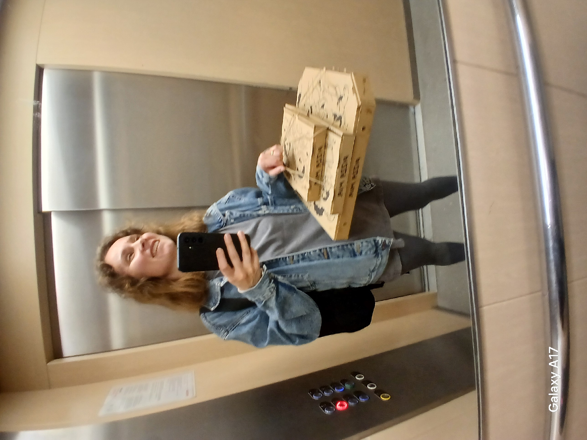An adult female is captured in a selfie reflected in a mirror-lined elevator in Yekaterinburg, Russia. She has medium-length curly brown hair and is smiling, looking towards the black smartphone she holds in her right hand. She wears a light grey top beneath a blue denim jacket and dark leggings, with a black backpack partially visible on her left shoulder. On her left ring finger, a ring is visible. Her left hand supports a stack of three brown cardboard pizza boxes, each featuring the "БУМ ПИЦЦА" (Boom Pizza) branding. The elevator interior consists of reflective silver-colored doors and light beige side panels. A control panel on the left wall contains multiple round buttons, with one red button illuminated, along with blue, green, black, and yellow buttons. A white document with Cyrillic text is posted on the beige wall. A chrome handrail runs horizontally across the lower portion of the reflective surface, and "Galaxy A17" is watermarked in the bottom-left corner of the image.