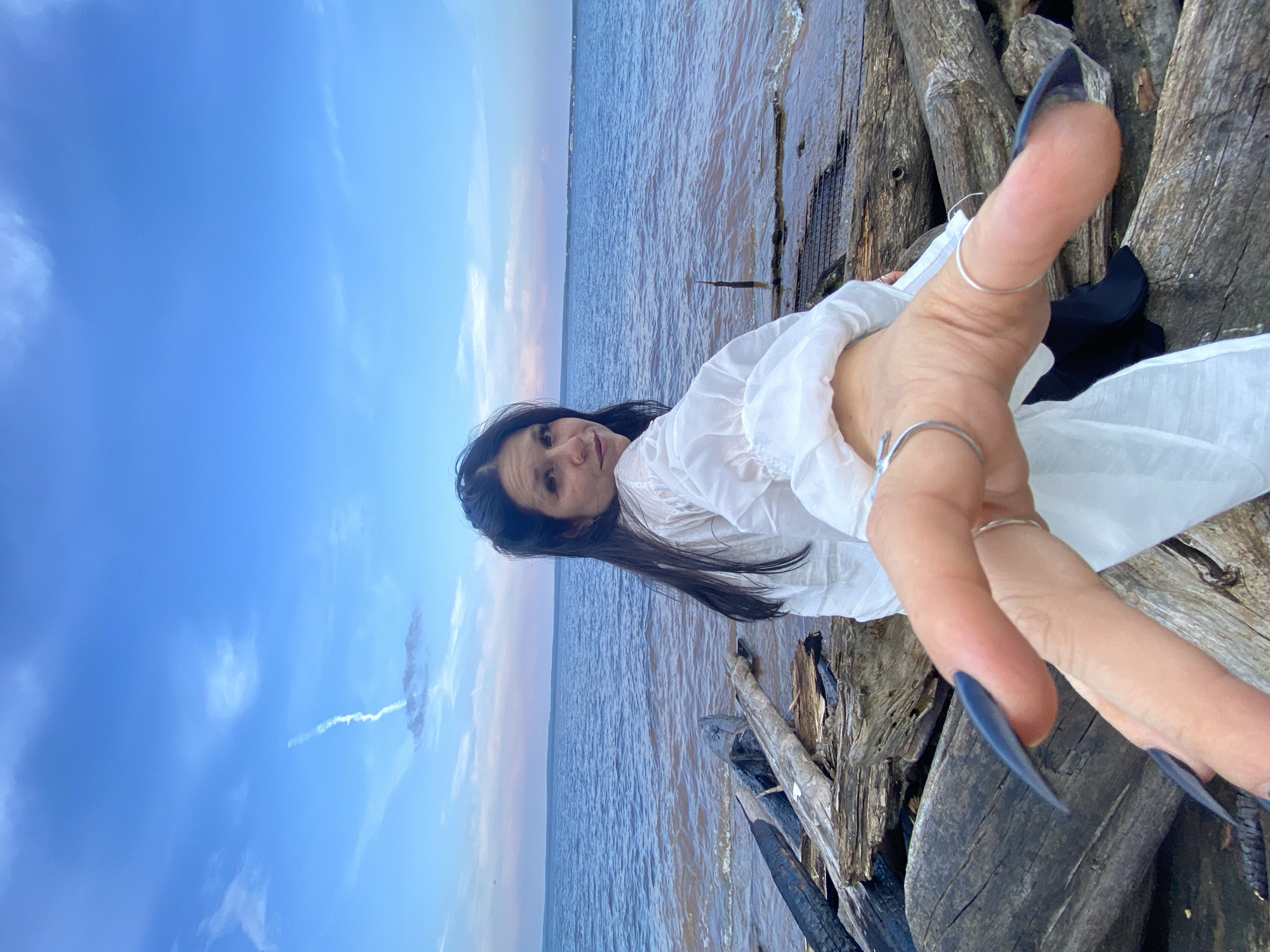 A woman with long, dark hair is positioned on a shore of weathered logs, facing away from the camera and looking over her shoulder. She is wearing a white, flowing top. Her right hand is extended towards the viewer, with long, dark blue fingernails and rings on her fingers. The background shows a calm body of water with a pinkish and blue sky overhead, dotted with wispy clouds and a contrail. The location is the Kama River embankment in Chaikovsky, Russia.