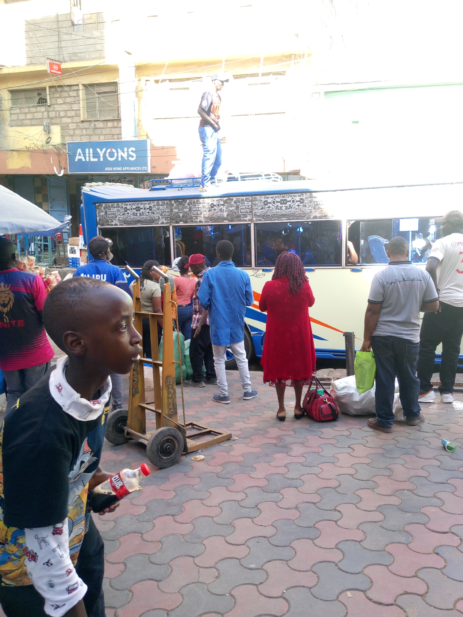 The image portrays a street scene in Nairobi, Kenya, characterized by bustling activity around a public transportation vehicle, likely a "matatu". A young boy stands in the foreground, holding a soda bottle. To the left of the child, individuals are seen standing and appearing to interact with the matatu, some seemingly boarding or waiting. A man stands on the roof of the matatu.
A store with the sign "AILYONS ADIA HOME APPLIANCES LTD" can be seen. The overall scene indicates a busy public space typical in urban environments in Nairobi. The ground is paved with interlocking concrete blocks.