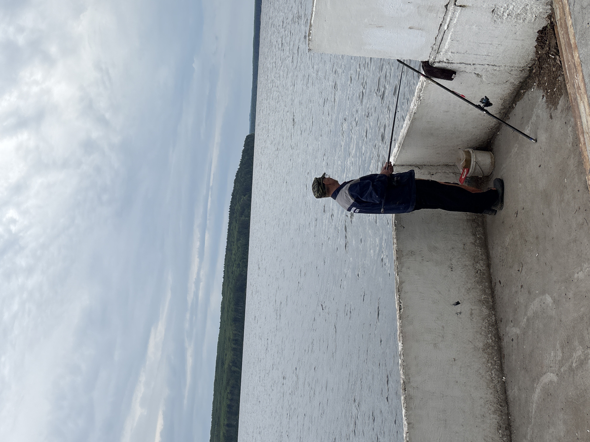 The image captures a daytime outdoor scene in Chaikovsky, Russia, featuring an individual fishing. An adult male, wearing a camouflage cap, a dark blue jacket with light grey sections, and dark trousers, stands on a sturdy concrete pier or embankment. He is holding a fishing rod, its line extending into the wide body of water before him. Another fishing rod is propped against the concrete wall to his right, next to a white bucket.
The water surface is textured with small ripples, suggesting a light breeze. In the distance, a long, dense treeline of dark green conifers stretches across the horizon, bordering the far side of the expansive waterway. The sky above is largely covered with light grey, scattered clouds, indicating overcast or partly cloudy weather. The concrete structure shows some wear and minor debris along its edges. The person appears focused on his fishing activity in a calm, natural setting. No discernible text is visible within the image.