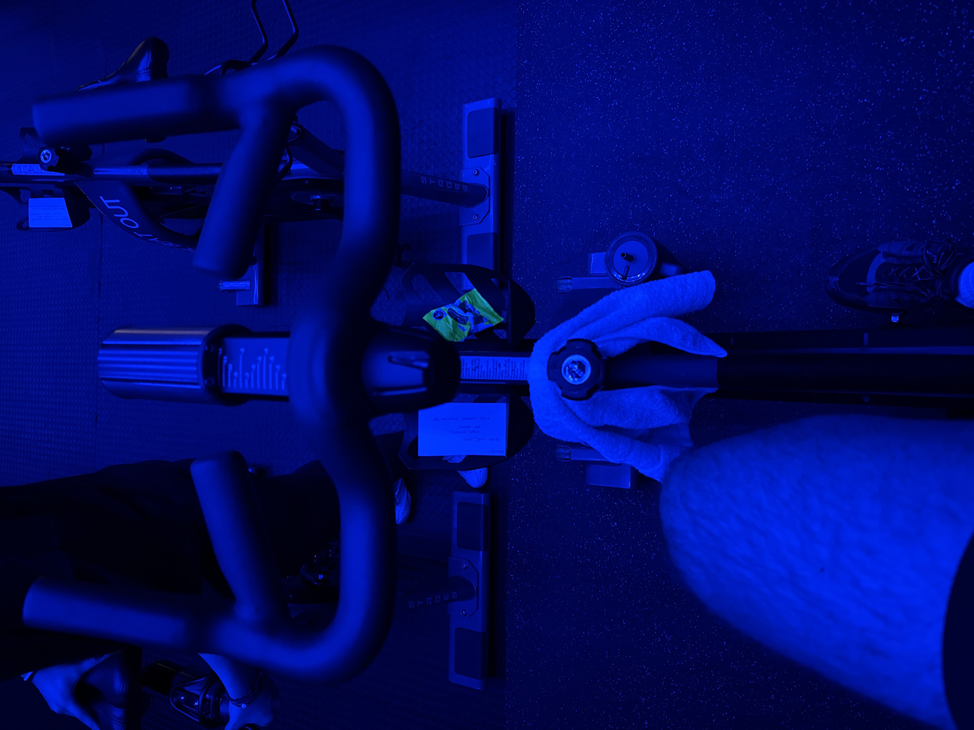 The image captures an overhead view of a person engaged in an indoor cycling session, bathed in a dominant, artificial blue light.
The primary subjects are parts of a person and a stationary exercise bike. On the right, a person's lower leg and foot, clad in a dark athletic shoe, are visible. A white towel is draped over the bike's frame near a water bottle or similar cylindrical container, suggesting the presence of hands just out of frame. The stationary bike itself features dark handlebars, a seat (partially visible), and a sturdy frame with various adjustment points and controls.
The setting is an indoor space, likely a gym or a home workout area, characterized by a dark, speckled or textured floor. The strong, uniform blue lighting creates a distinct, almost ethereal atmosphere, commonly found in spin studios or specialized workout rooms, and suggests either a specific mood lighting setup or a nighttime setting.
The activity taking place is a workout session on the stationary bike. The towel and water bottle indicate that the person is actively exercising or has just completed an intense session.
Notable details include:
* The pervasive blue light which colors all elements.
* A small, bright yellow and green packet, possibly a snack or supplement, rests on the bike's frame.
* A small white rectangular note with handwritten text is affixed to the bike frame, just below the handlebars.
Visible text includes:
* "BEAT OUT" written in white on the bike's frame on the left side.
* "STRADDLE" (or similar) is vertically aligned on the bike's central adjustment post.
* On the white note, faint and partially obscured handwritten text includes "Workout plan" and "Calories," along with other illegible words and numbers.