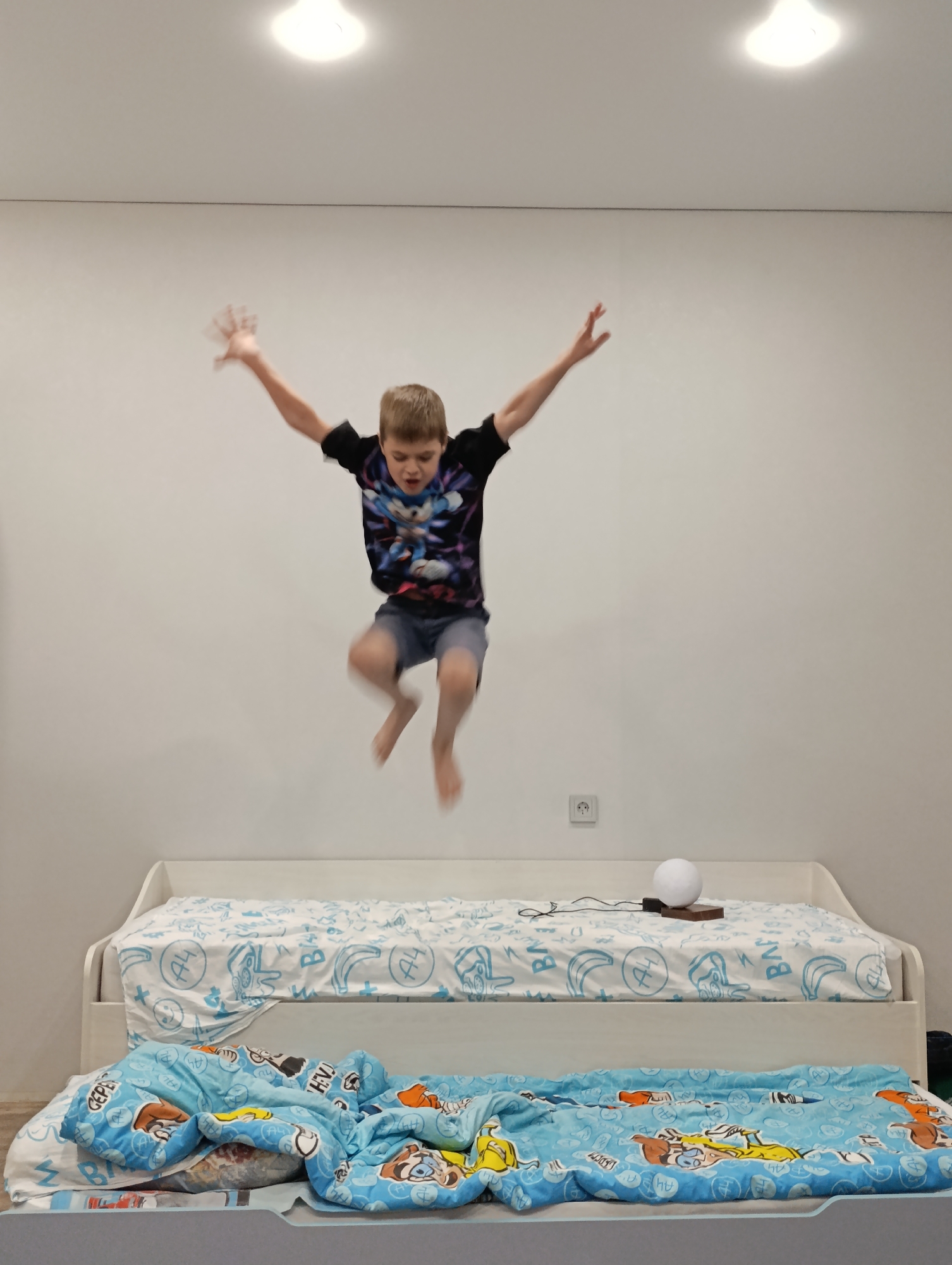 A young boy is captured mid-air, seemingly jumping above a bed. He wears a black t-shirt featuring a graphic print, and dark shorts. His arms are extended outwards and upwards. The background consists of a plain white wall. Below the boy is a bed covered with a white sheet patterned with blue graphics. A smaller bed underneath is covered with a blue blanket. A small round lamp rests on the first bed. A electrical outlet can be seen on the wall. The location is Chaikovsky, Russia.