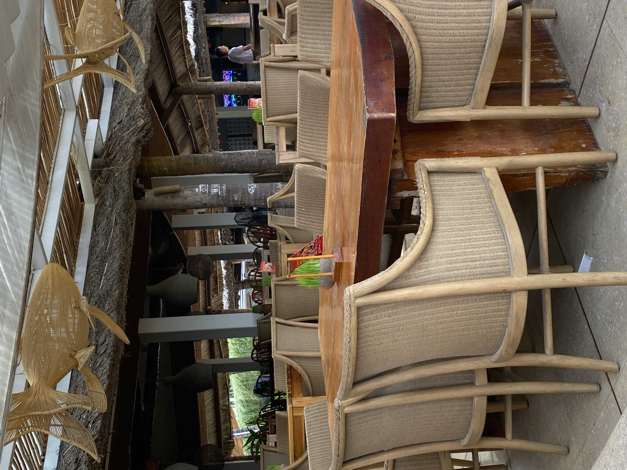 The image depicts an outdoor dining area, likely a restaurant or cafe, in Nha Trang, Vietnam. The setting features a thatched roof structure supported by thick wooden beams and palm tree trunks, creating a tropical and rustic ambiance. Several tables are visible, with chairs made of woven rattan. One table in the foreground is a long, wooden surface with a curved edge. On this table, a small planter with green sprouts is placed, along with a red menu or sign. The scene is illuminated by natural light, suggesting it's daytime.
In the background, through an opening in the structure, glimpses of lush greenery and more tables and chairs can be seen. A few people are faintly visible in the distance, including one person standing near some screens displaying colorful images. The overall atmosphere appears relaxed and inviting, typical of a seaside resort destination. The distinctive woven fish decoration hanging from the ceiling adds a unique artistic touch to the decor. The image is oriented vertically, suggesting it was taken with a phone camera.